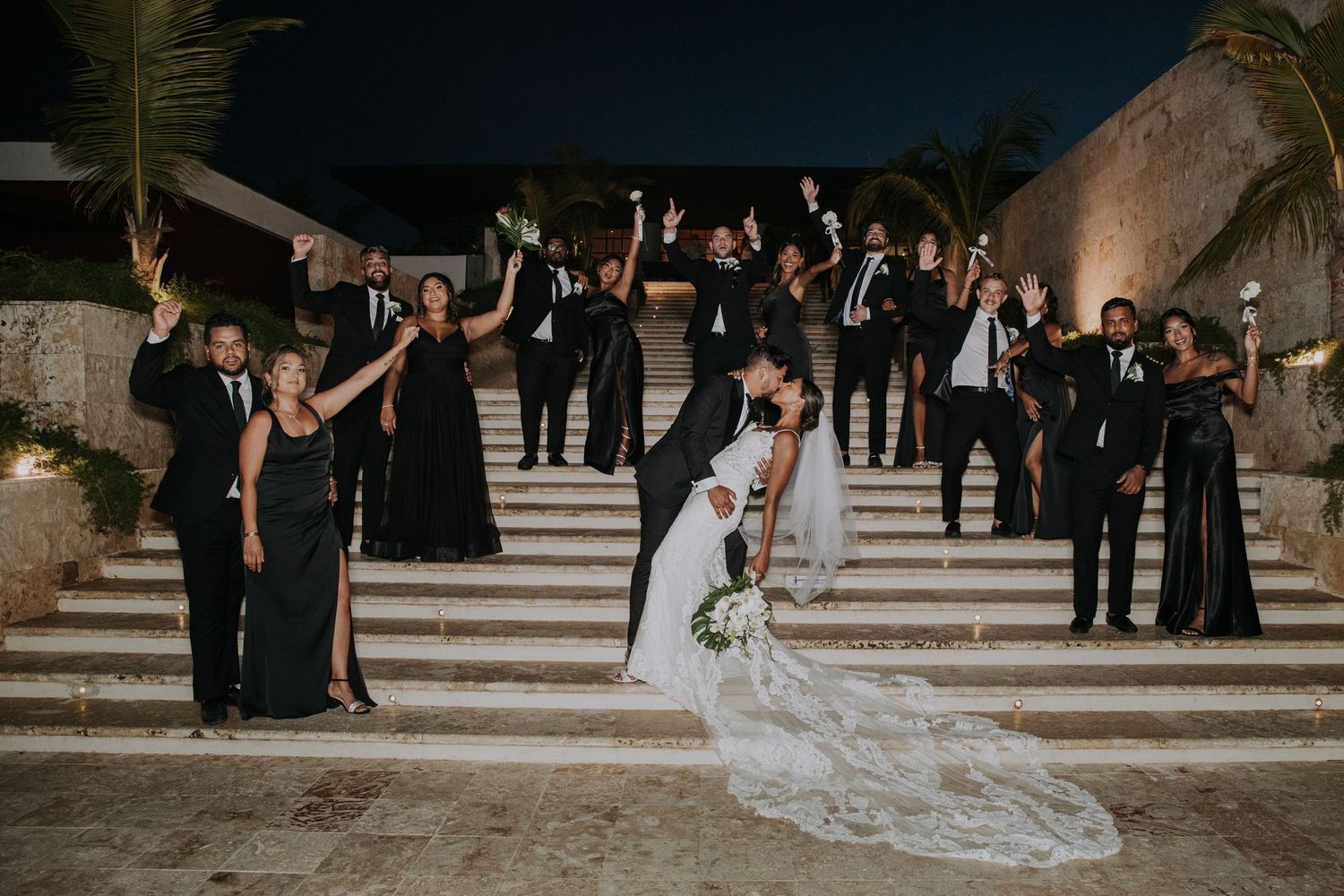 Wedding guests in black attire gather around a couple on grand stone steps.