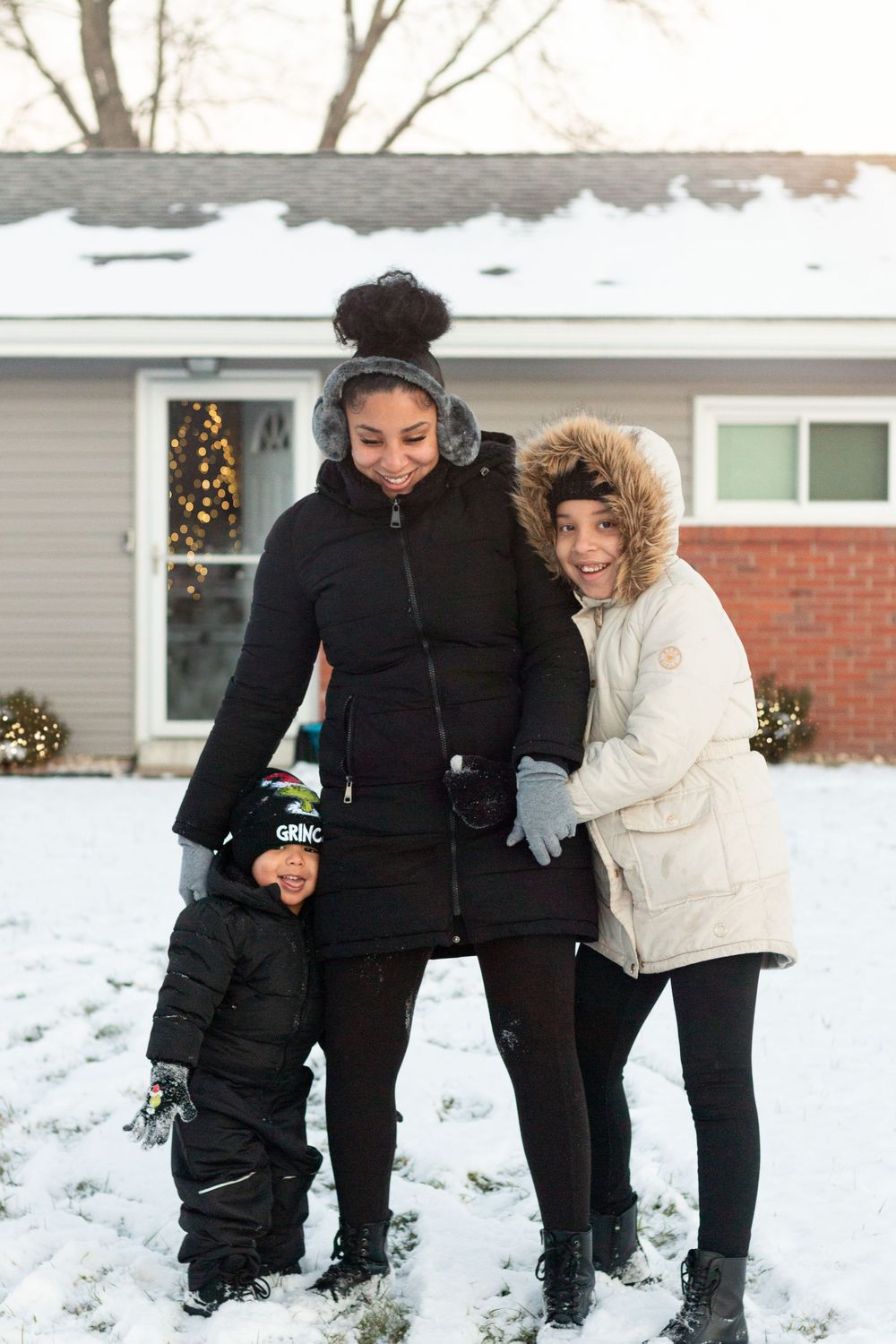 Family wearing winter coats stands together in snowy front yard with holiday lights visible in background.