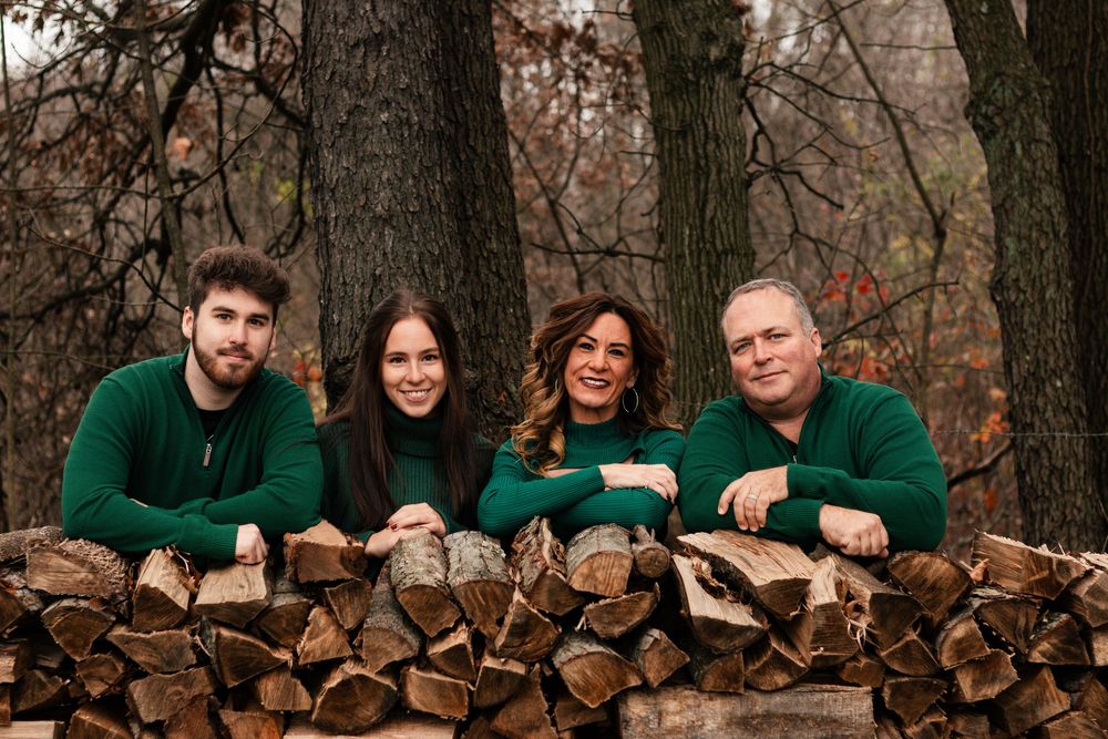 A group poses behind a rustic wood pile in an autumn forest setting wearing coordinated green and brown outfits.