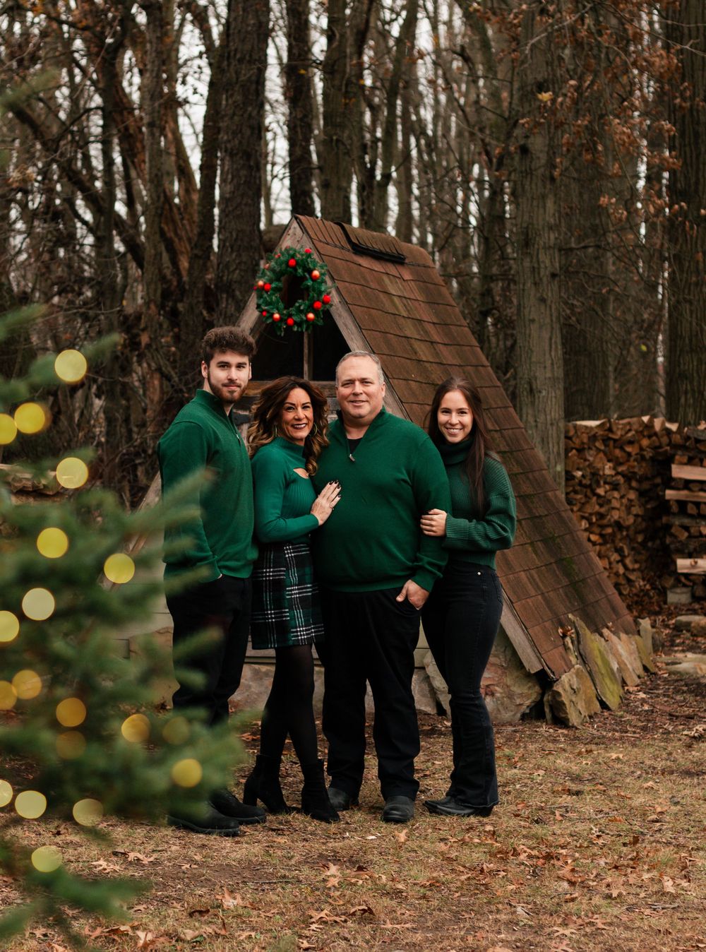 A family wearing matching green sweaters poses together in a wooded outdoor setting during autumn.