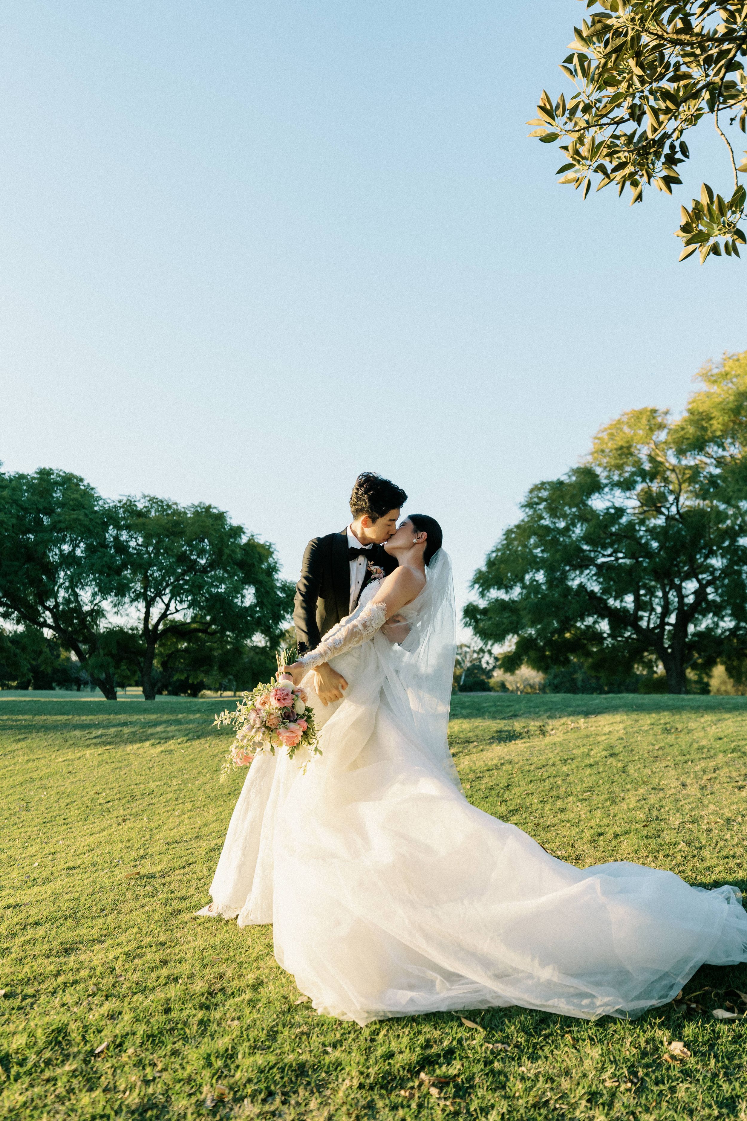 A couple embraces in a romantic outdoor wedding photo on a grassy hill during golden hour with trees in the background.
