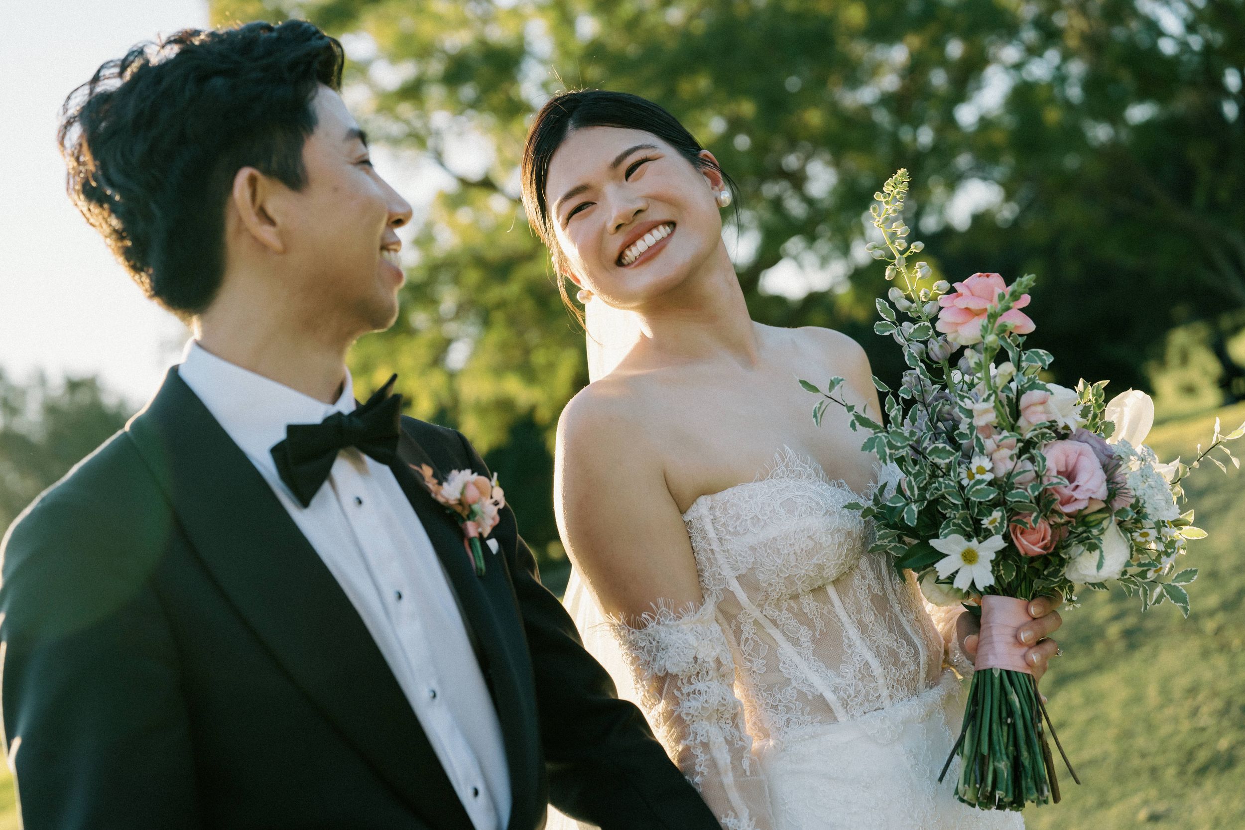 A couple in wedding attire share a joyful moment outdoors holding a soft pink and green floral bouquet.