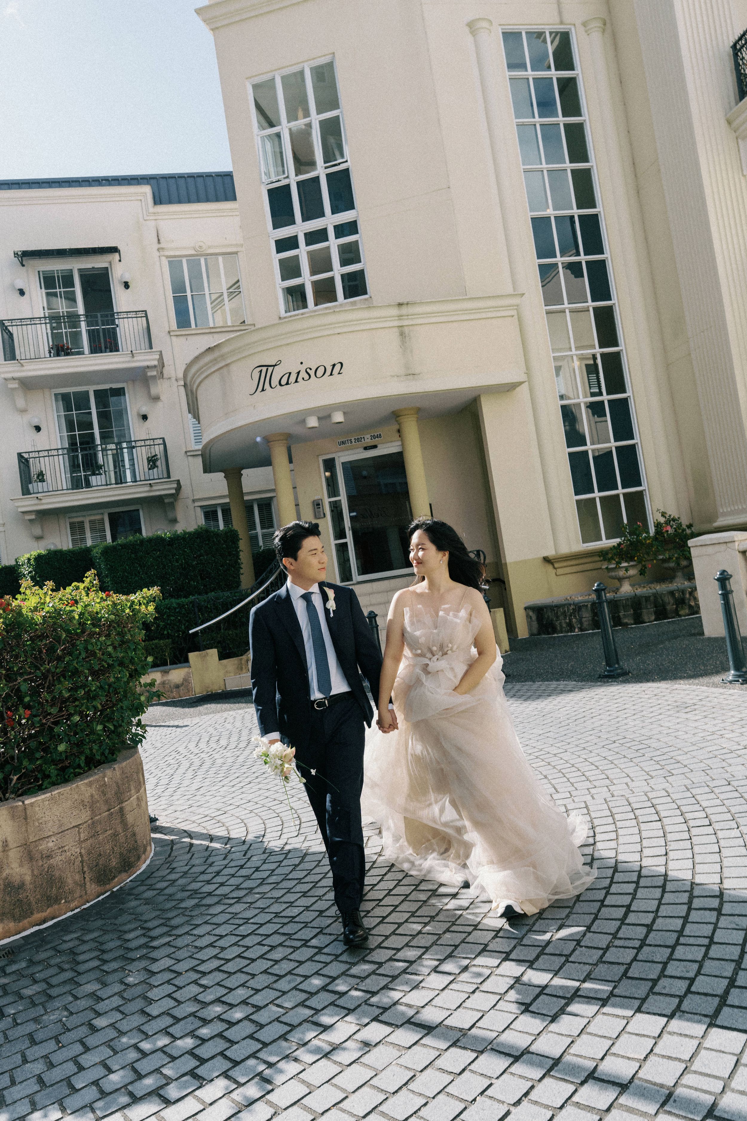 A couple in wedding attire walks along a curved brick path outside an elegant white building with classical architecture.