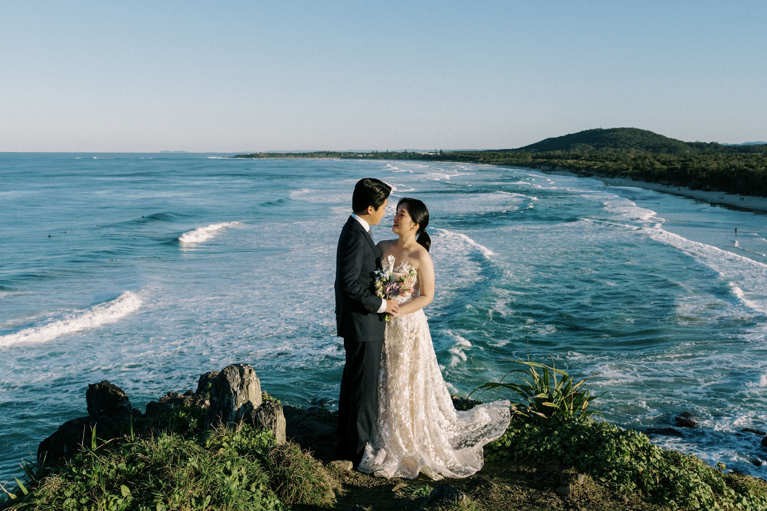 A couple admires the stunning coastal views from a scenic cliff overlooking turquoise ocean waves breaking on the shore.
