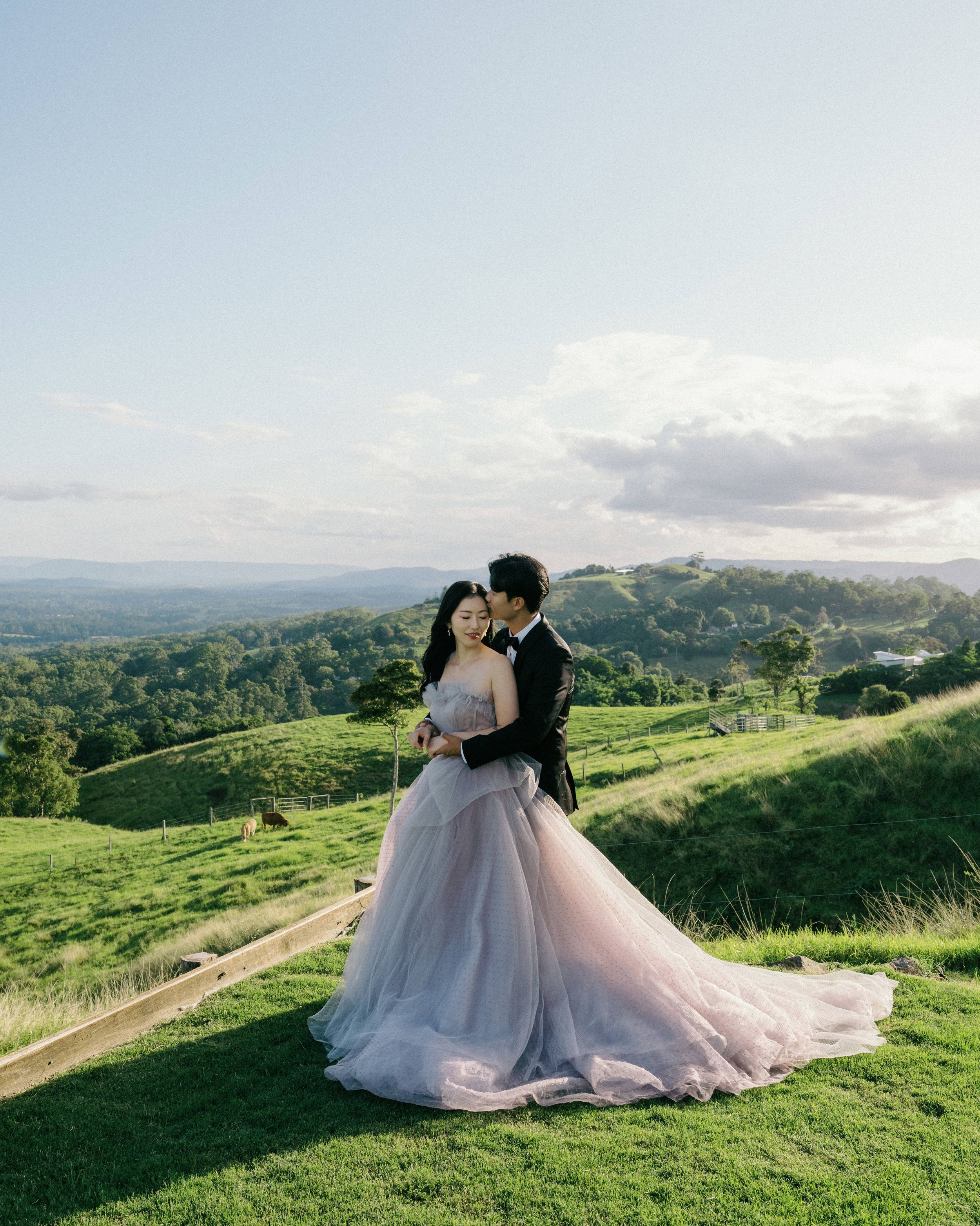 A couple in wedding attire share a romantic moment on a hilltop with rolling green hills and mountains in the background.