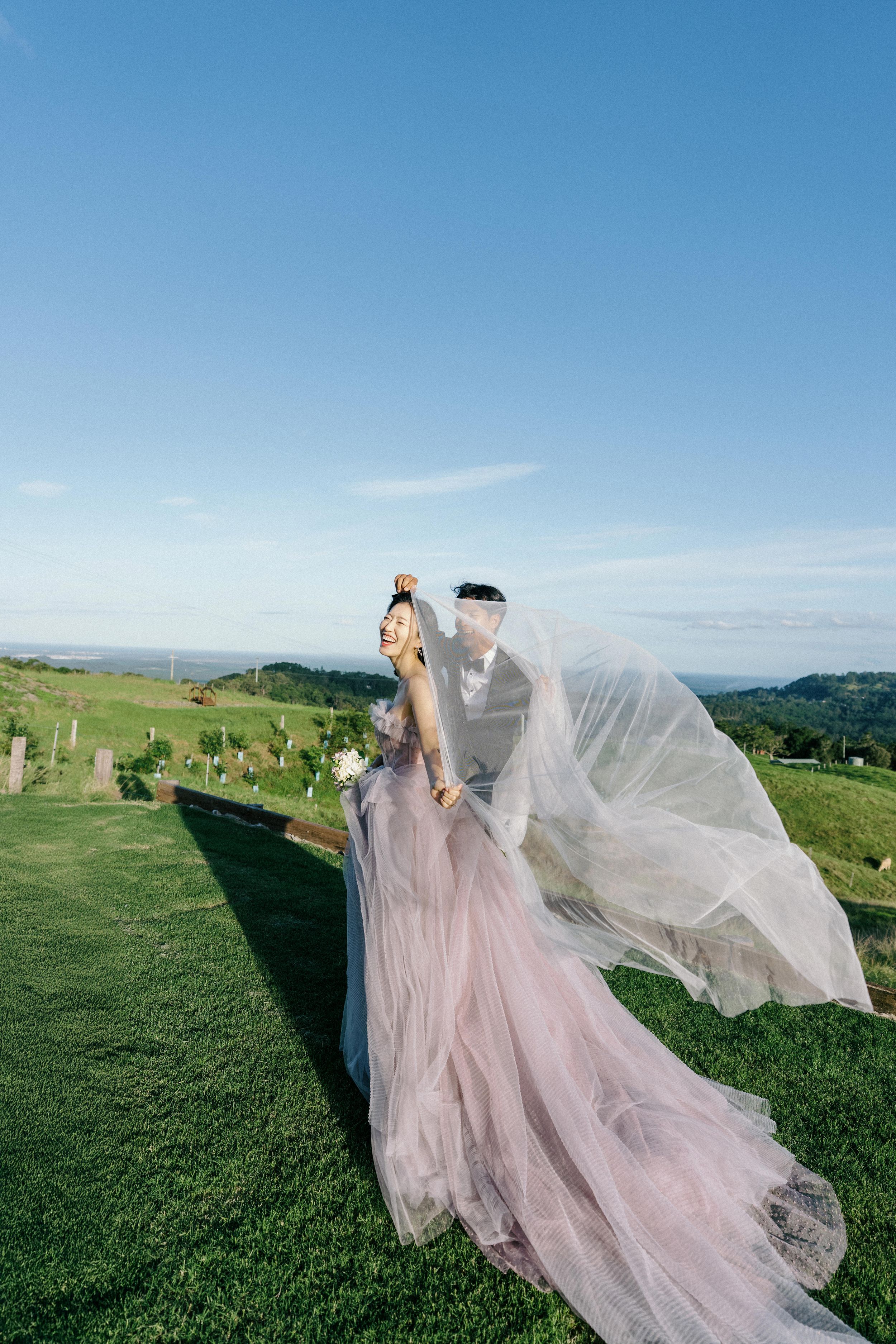 A wedding couple on a grassy hilltop with flowing pink tulle dress and veil against blue sky and mountain backdrop.