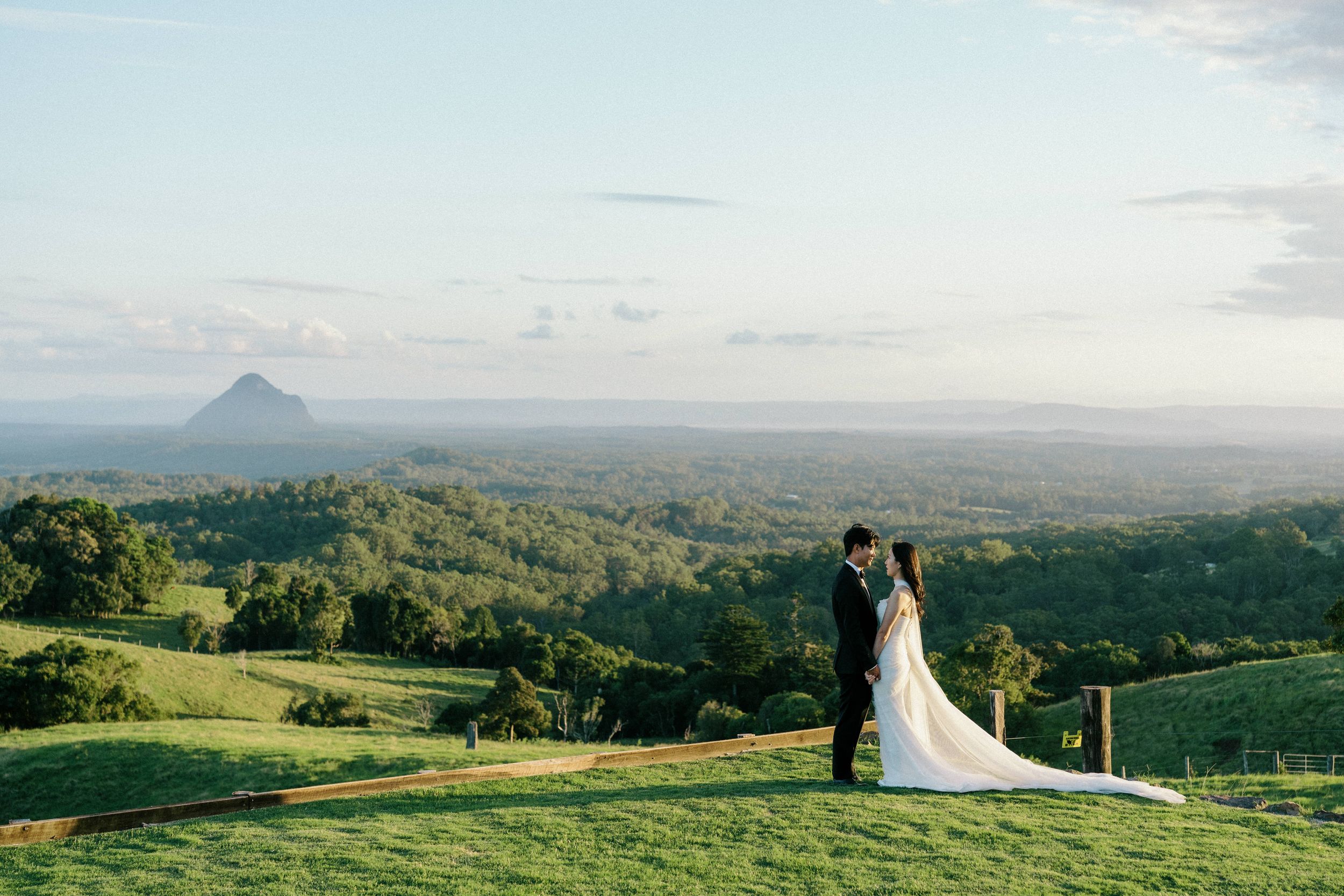 A couple stands on a hilltop overlooking lush green rolling hills and mountains in the distance at sunset.