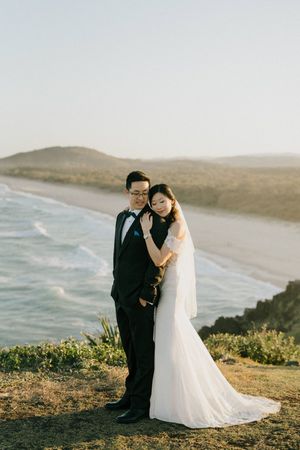 Couple in wedding attire stands on coastal clifftop overlooking a scenic beach at sunset.