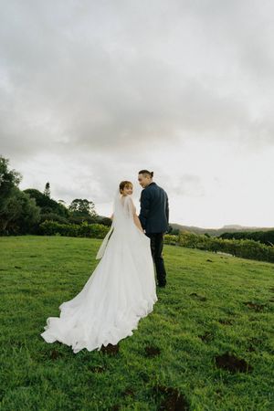 A couple stands together on a lush green hillside under cloudy skies with the bride wearing a flowing white wedding gown.