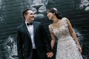 A bride in a beaded gown and groom in tuxedo share a joyful moment against a black and white graffiti wall.