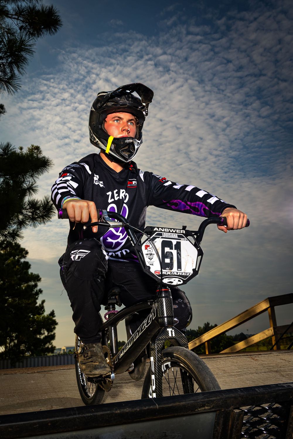 BMX rider in black and purple gear performs trick on ramp against cloudy sunset sky.