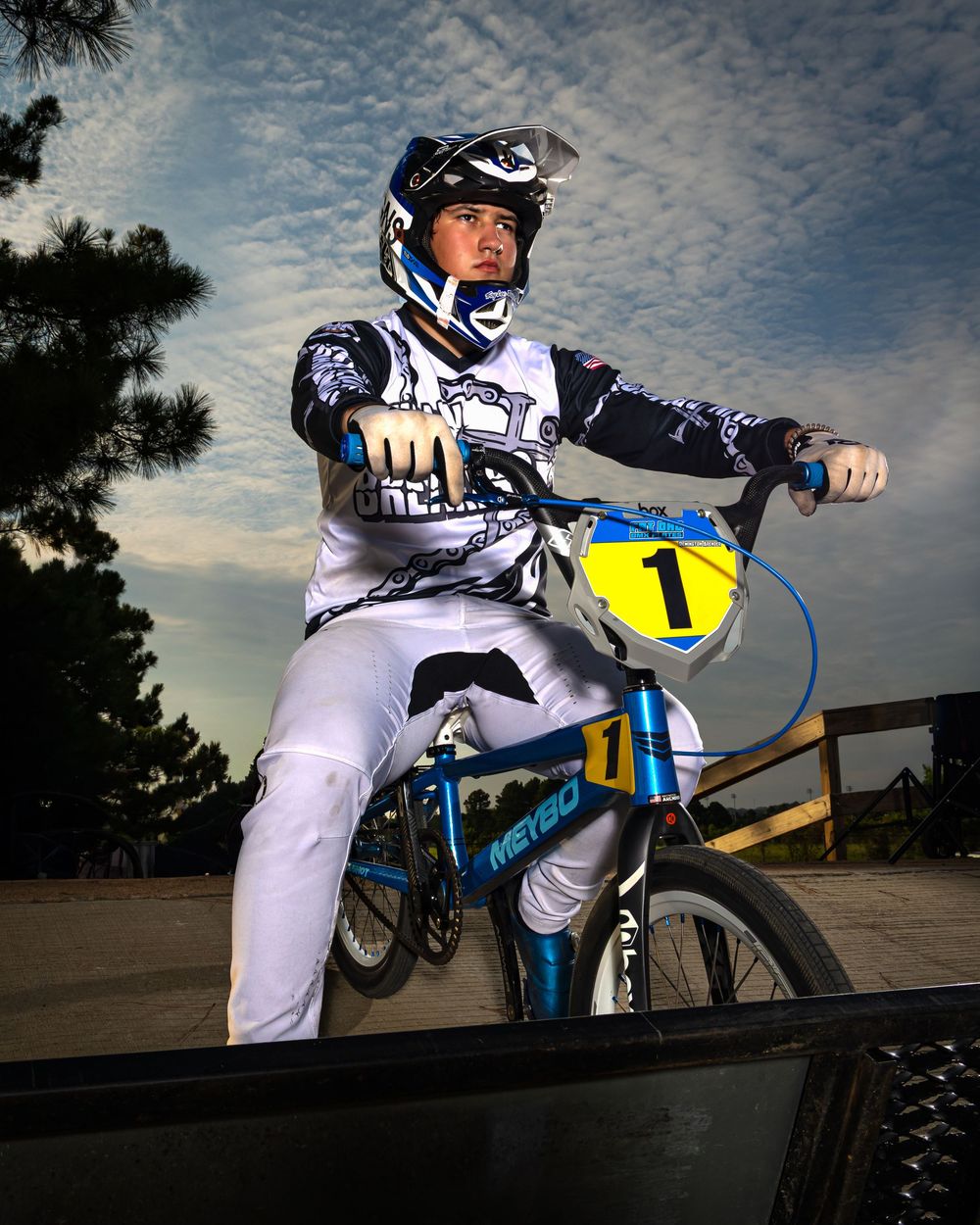 A BMX rider in white racing gear with number 1 plate poses on a blue bike against a cloudy sunset sky.