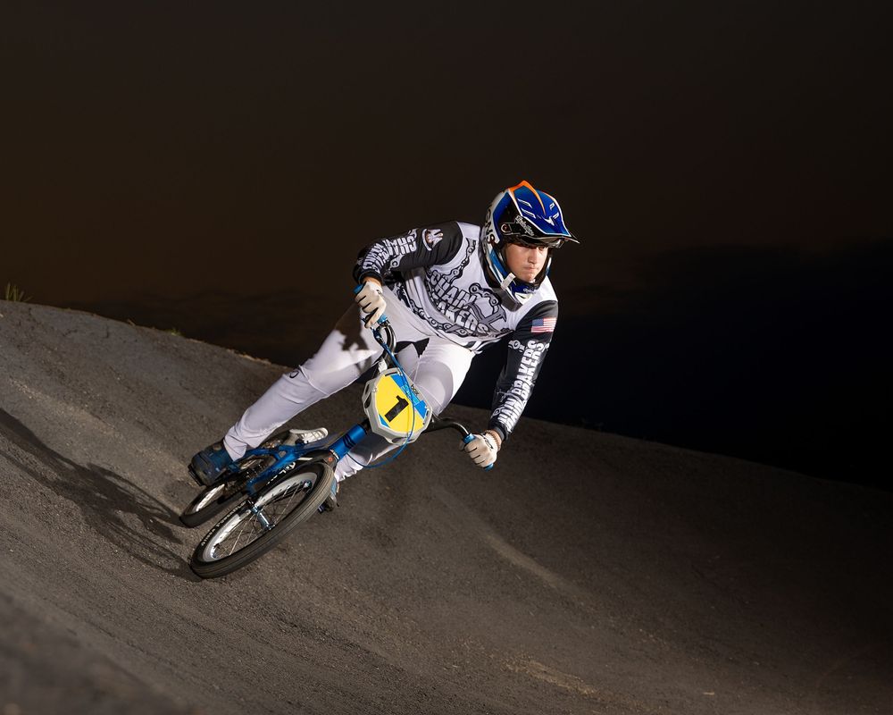 BMX rider in white jersey and blue helmet races down a dark dirt track at night.
