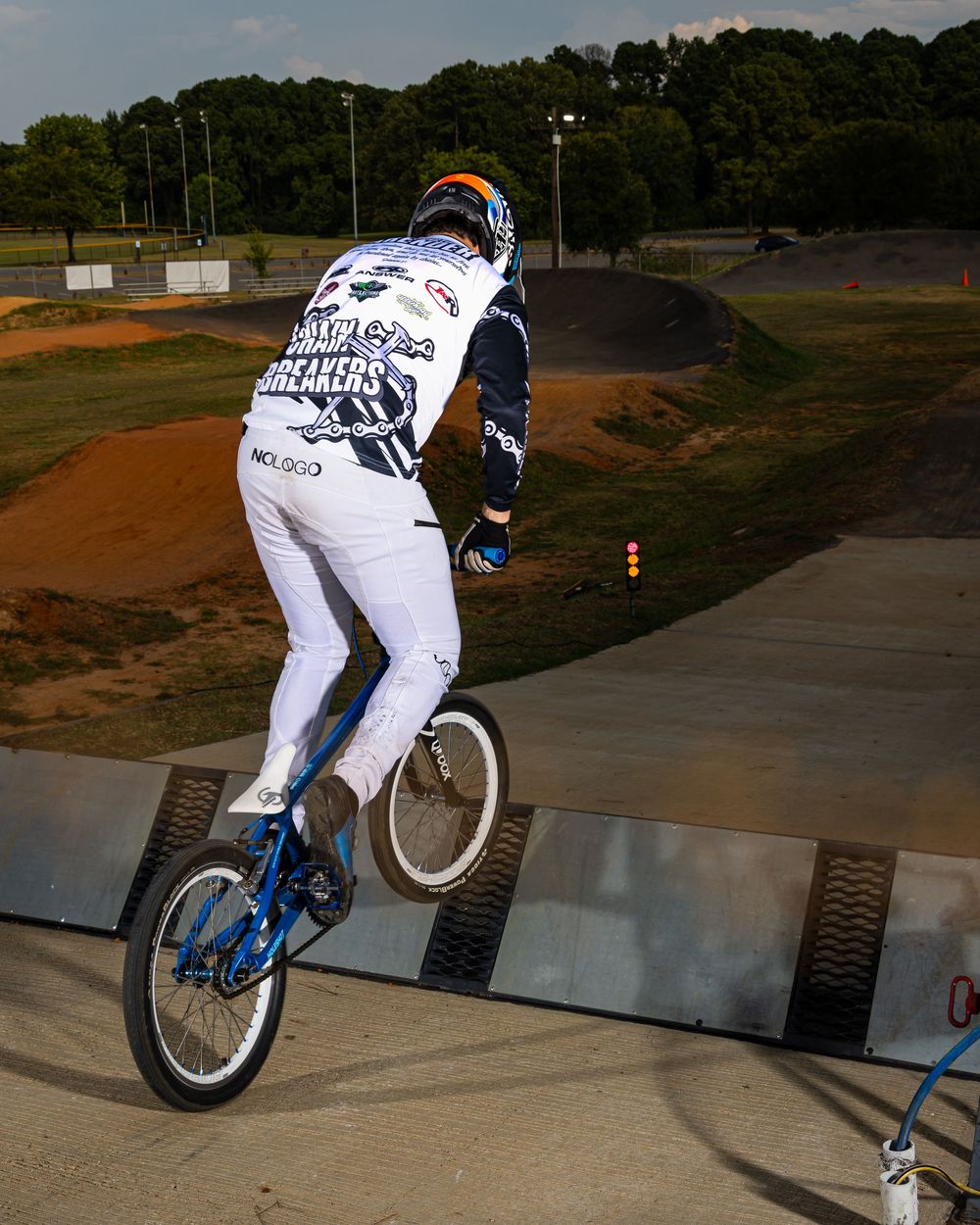 BMX rider in white uniform performing jump on blue bike at dirt track with ramp and trees in background.
