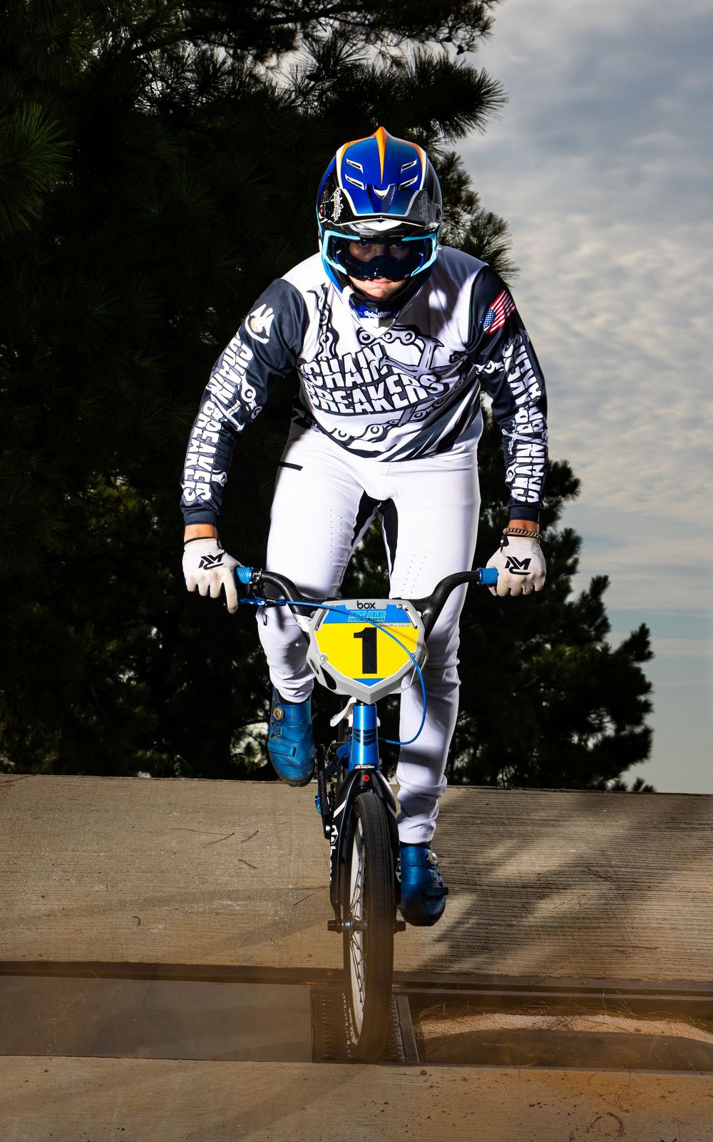 BMX rider in white and blue gear performs an aerial trick on a dirt track under cloudy skies.