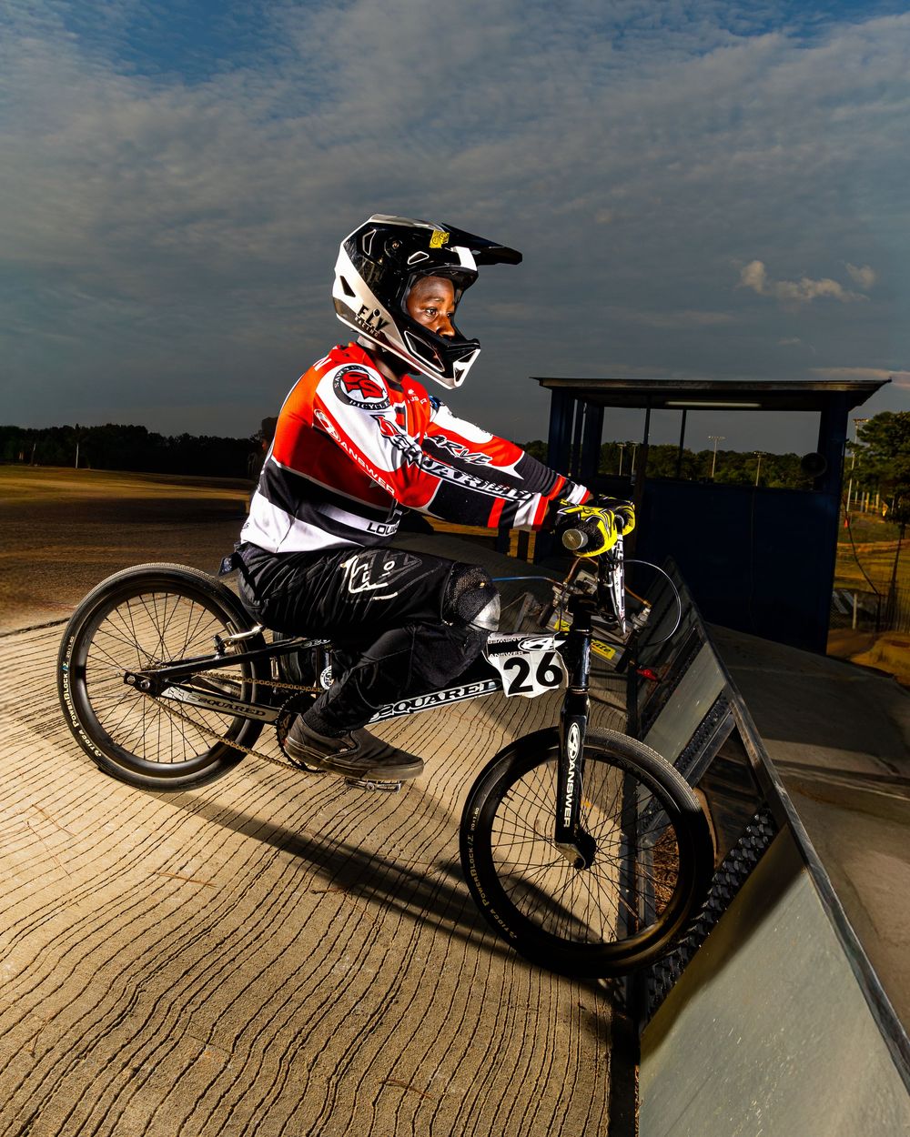 BMX racer on wooden ramp wearing helmet and racing gear at outdoor track during sunset.