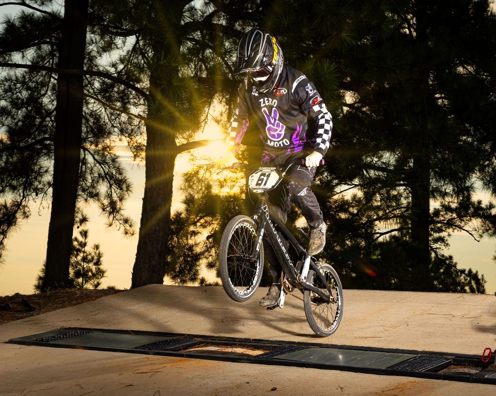 BMX rider performing jump on dirt track at sunset with sunburst through trees in background.