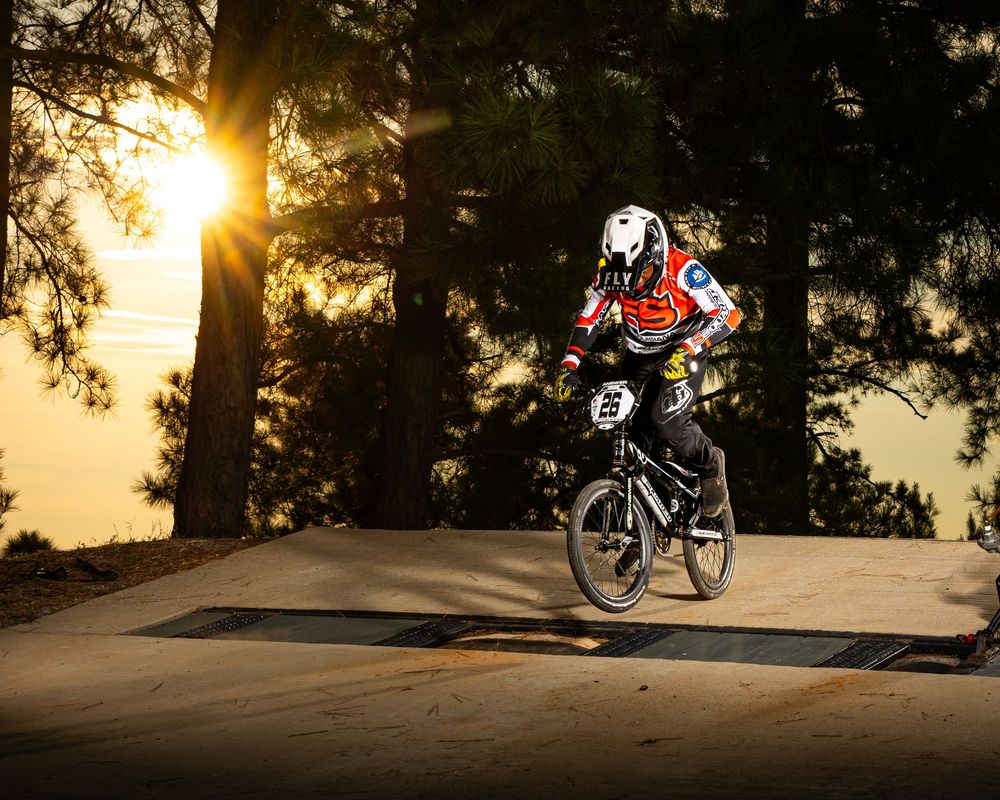 Mountain biker rides trail at sunset through silhouetted pine trees casting long shadows on winding dirt path.