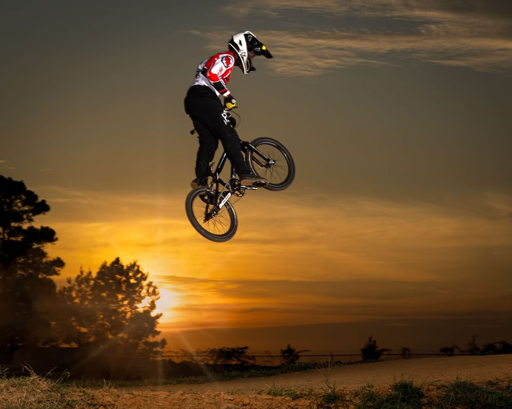 Silhouetted BMX rider performs aerial tricks against a dramatic orange sunset sky during a dirt jump session.