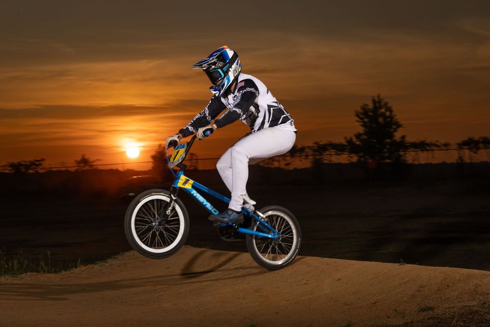 A BMX rider performs aerial tricks against a vibrant orange sunset sky on a dirt track.