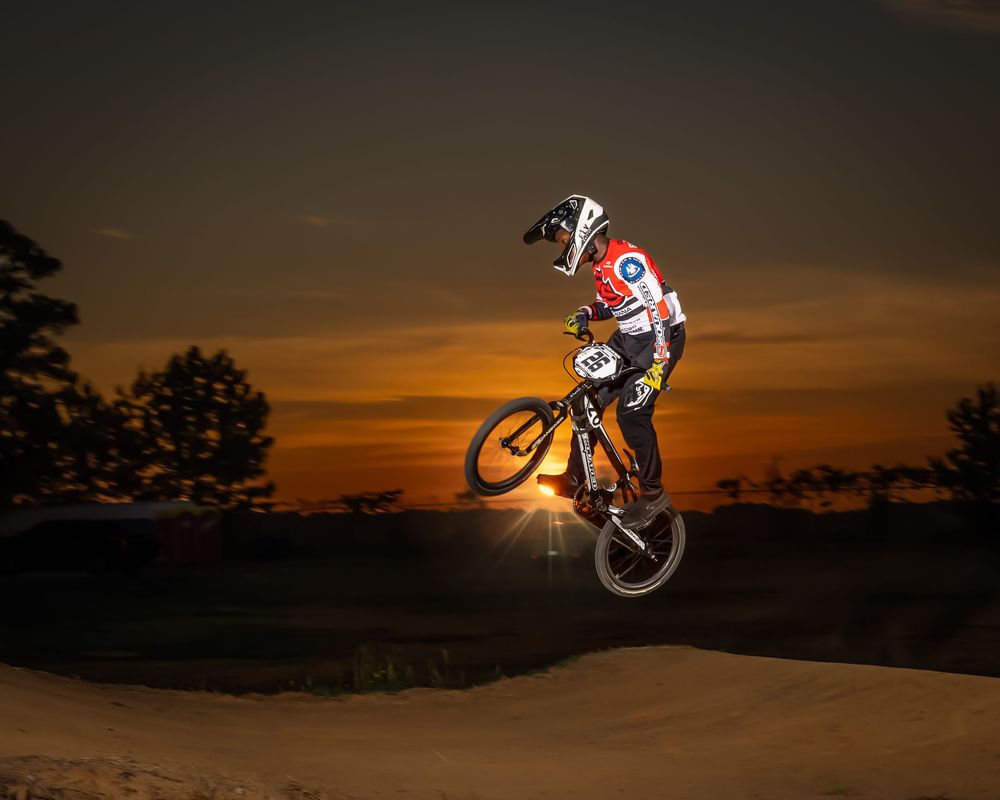Silhouetted BMX rider performs aerial trick against dramatic orange sunset sky over dirt jump track.