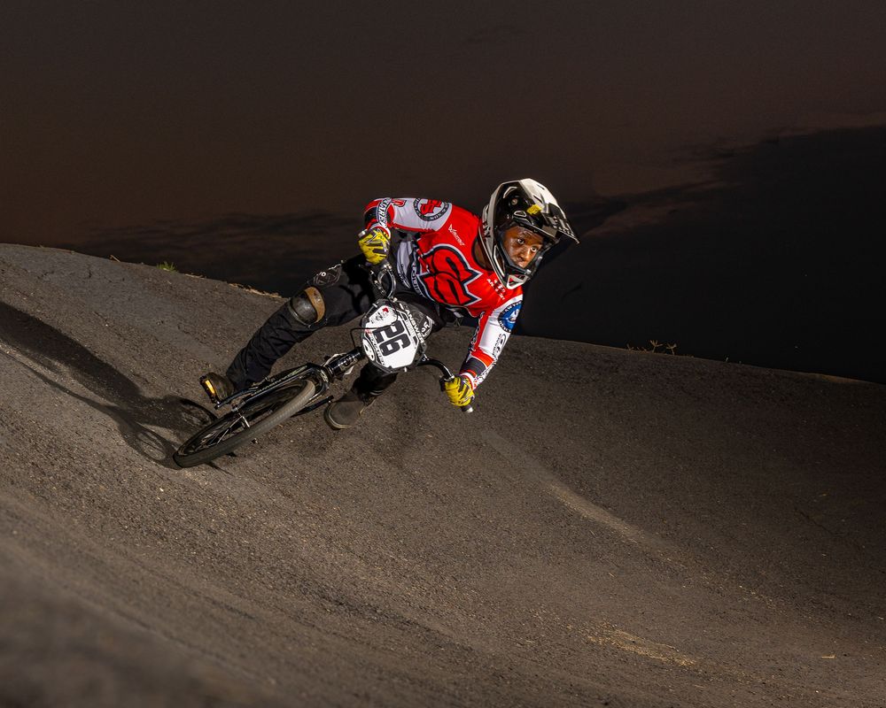 A motocross rider races down a steep dirt track in darkness, illuminated by spotlights during a night competition.