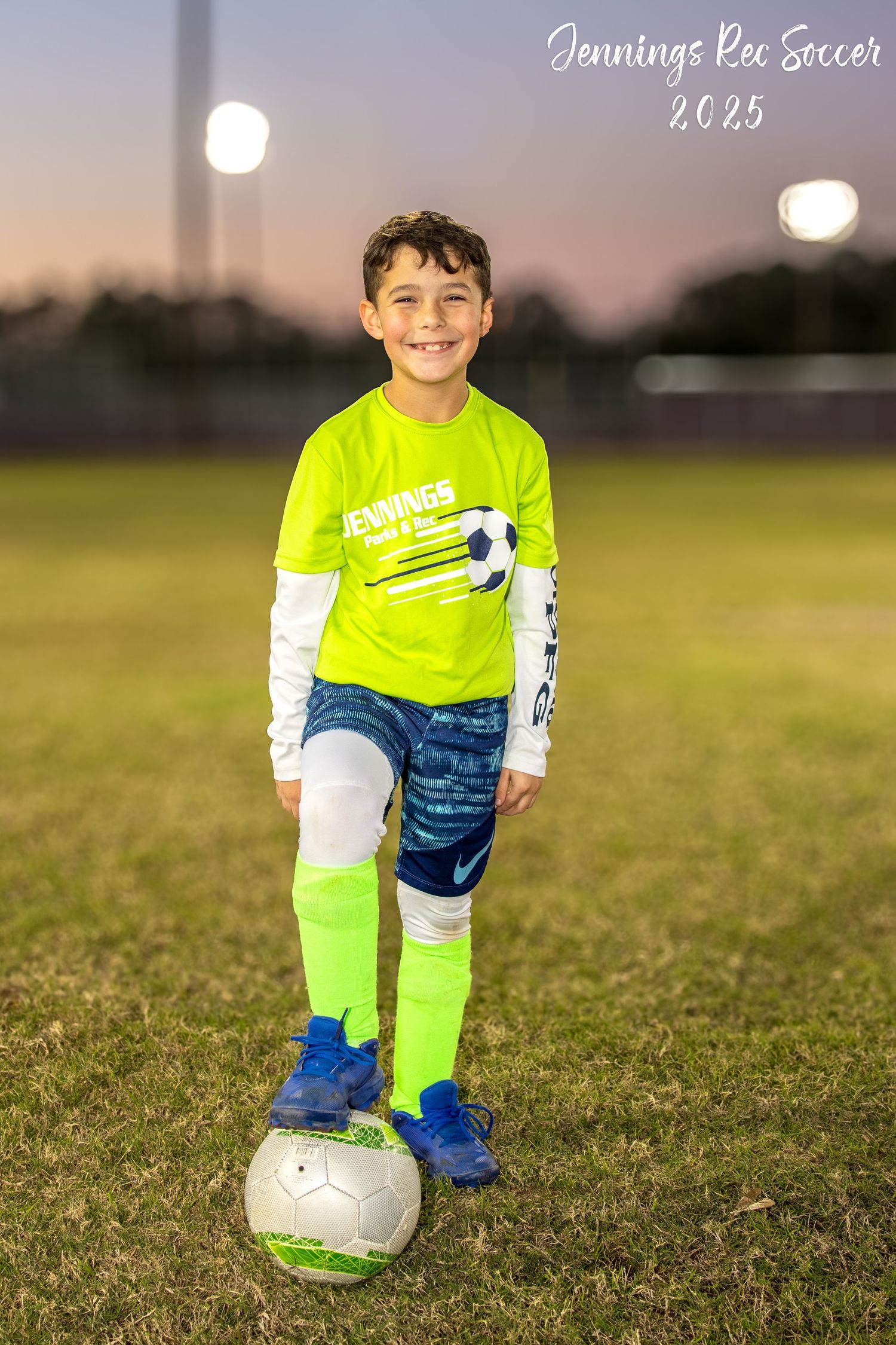 Young soccer player in neon yellow jersey and blue pants poses with ball on grass field at dusk.