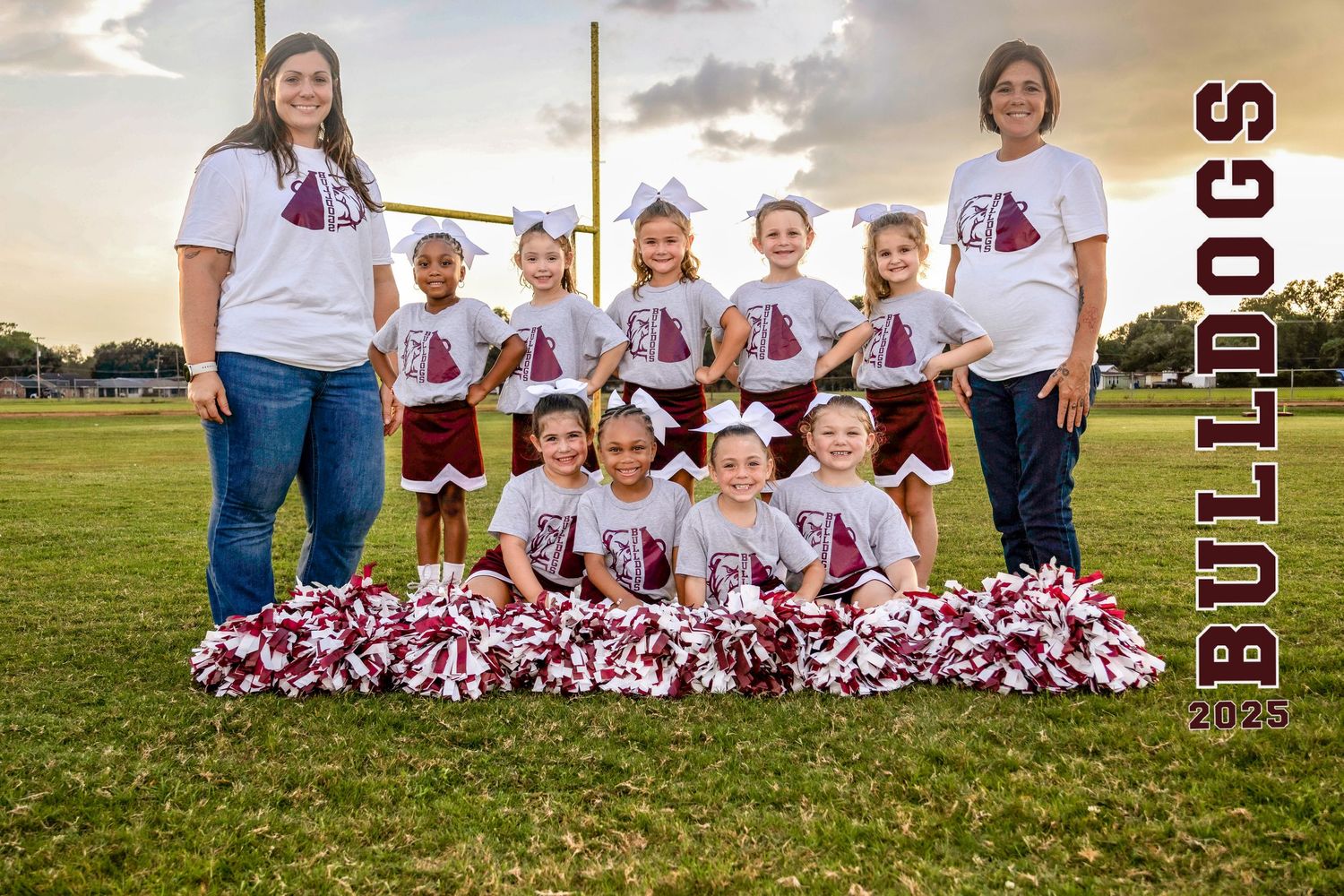 Youth cheerleading team poses for group photo on grass field with pom poms and matching uniforms at sunset.