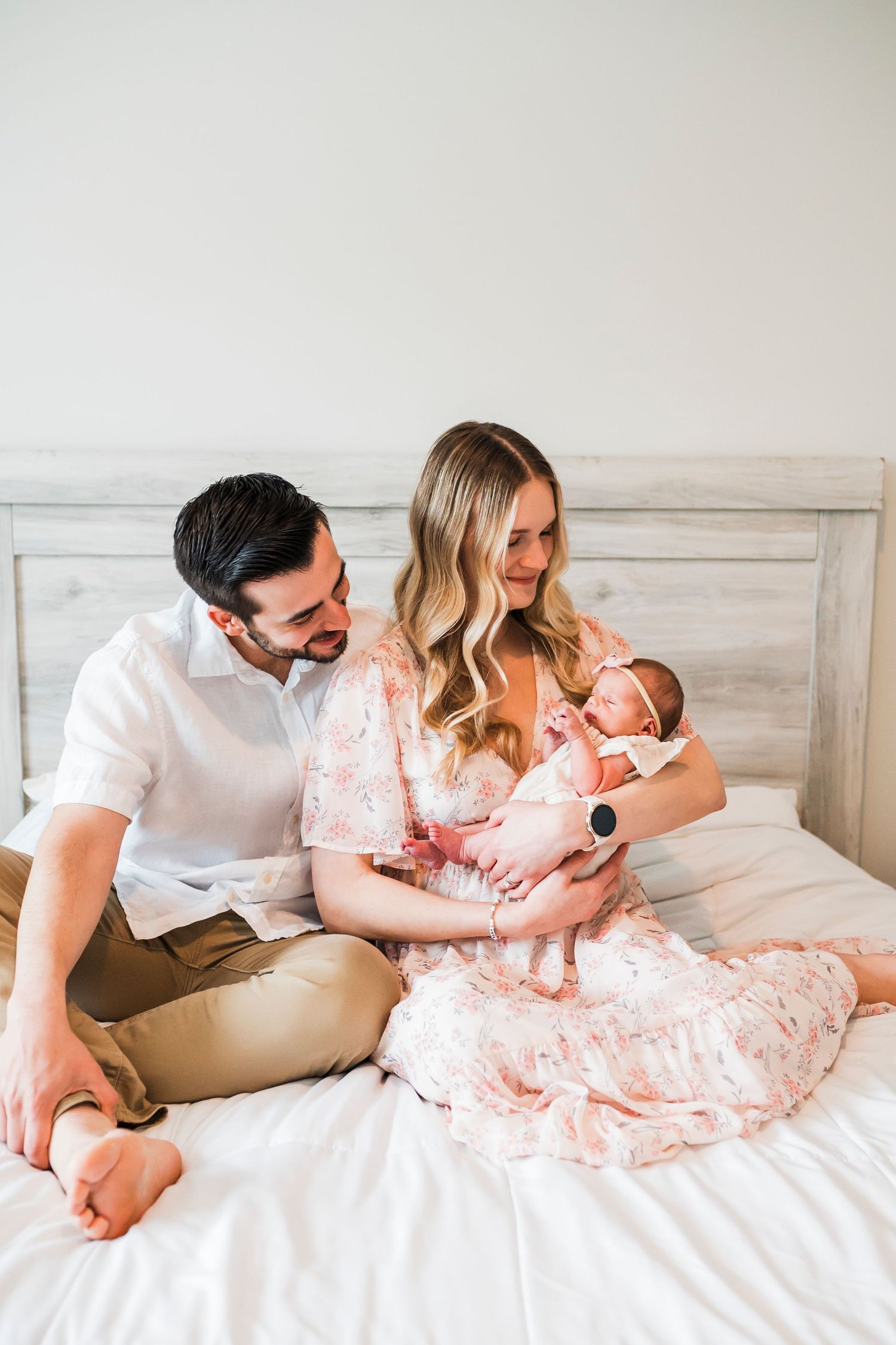 A couple relaxes on a white bed while bonding with their newborn baby in a light and airy setting.