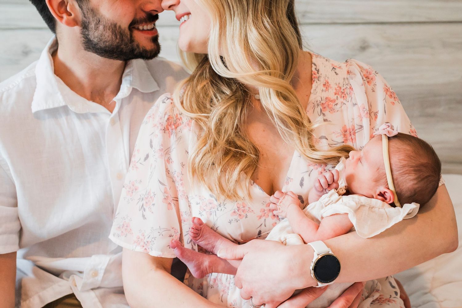 Parents wearing white and floral clothing share intimate moments with their newborn baby on a white bed.
