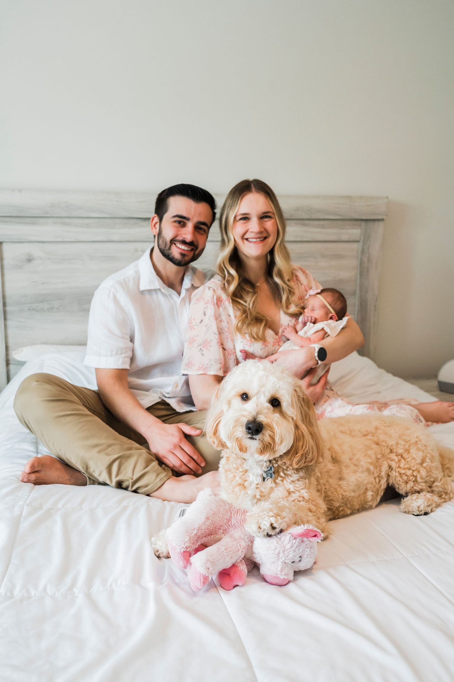 A family lounges on a white bed with their fluffy goldendoodle dog in a bright, airy bedroom setting.