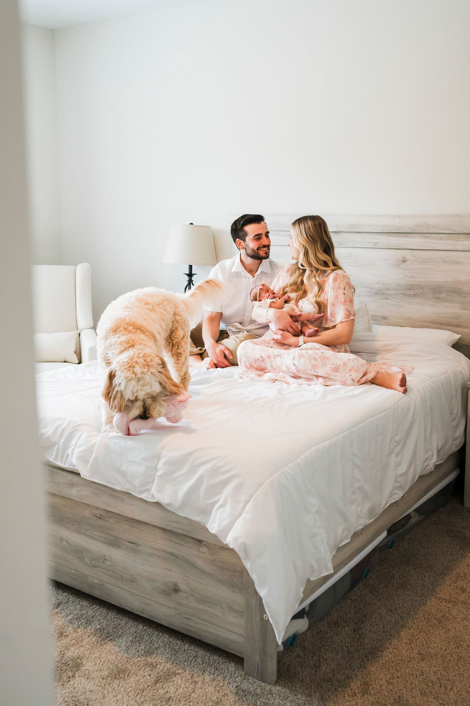 A couple relaxes on a white bed with their dog in a bright, minimalist bedroom setting.