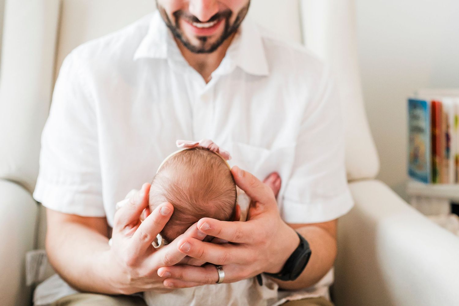 A person in a white shirt cradling a sleeping newborn baby while sitting indoors beside a bookshelf.