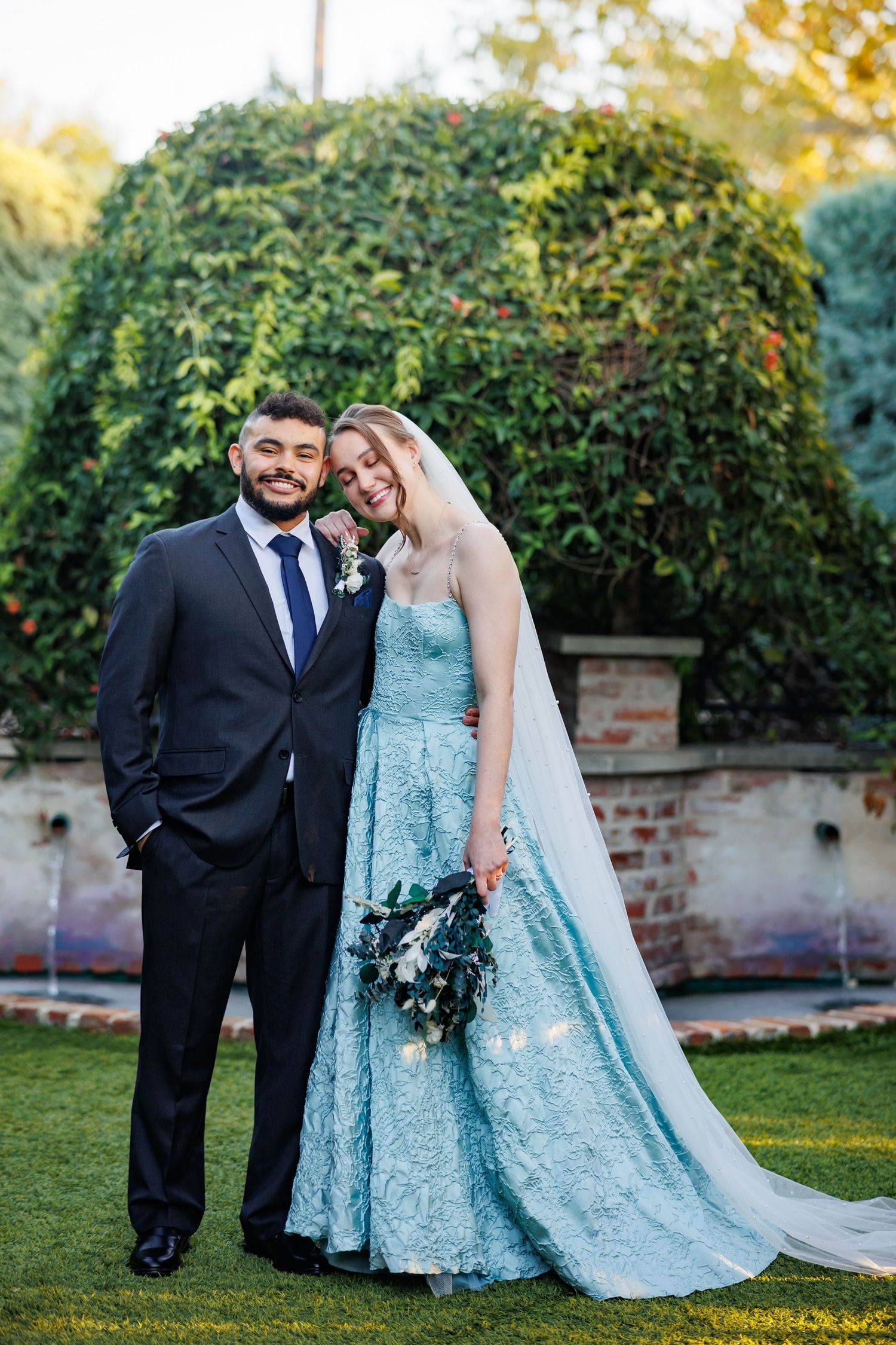 Wedding couple poses in garden wearing light blue lace gown with long veil and black suit with blue tie.
