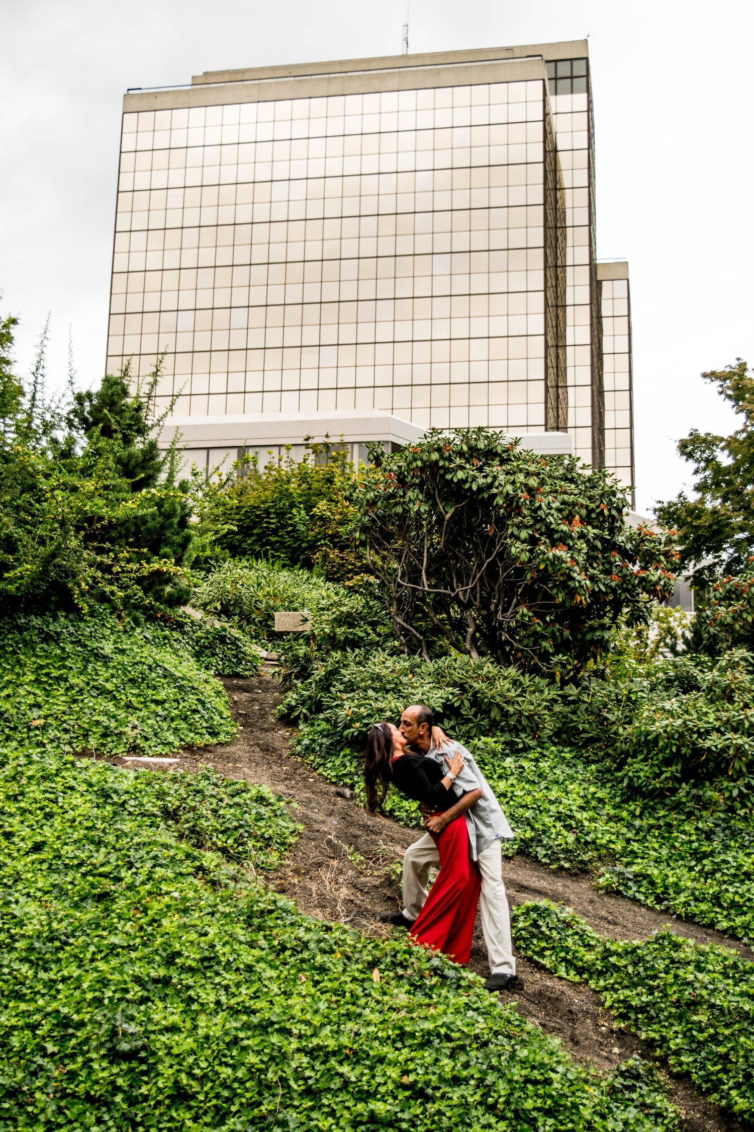 A couple in red and white outfits sharing a romantic moment near a wall with green foliage.