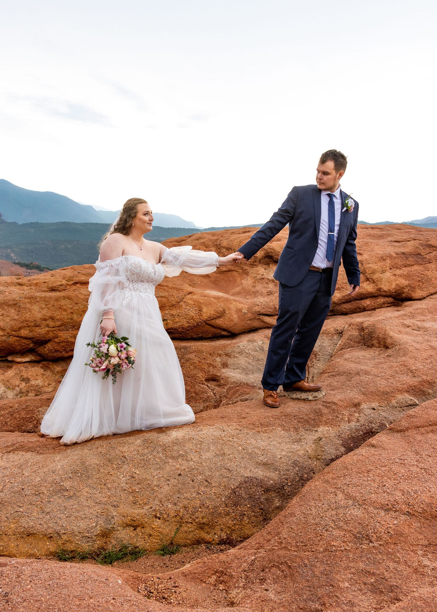Wedding couple posing playfully on red rock formations with mountain views.