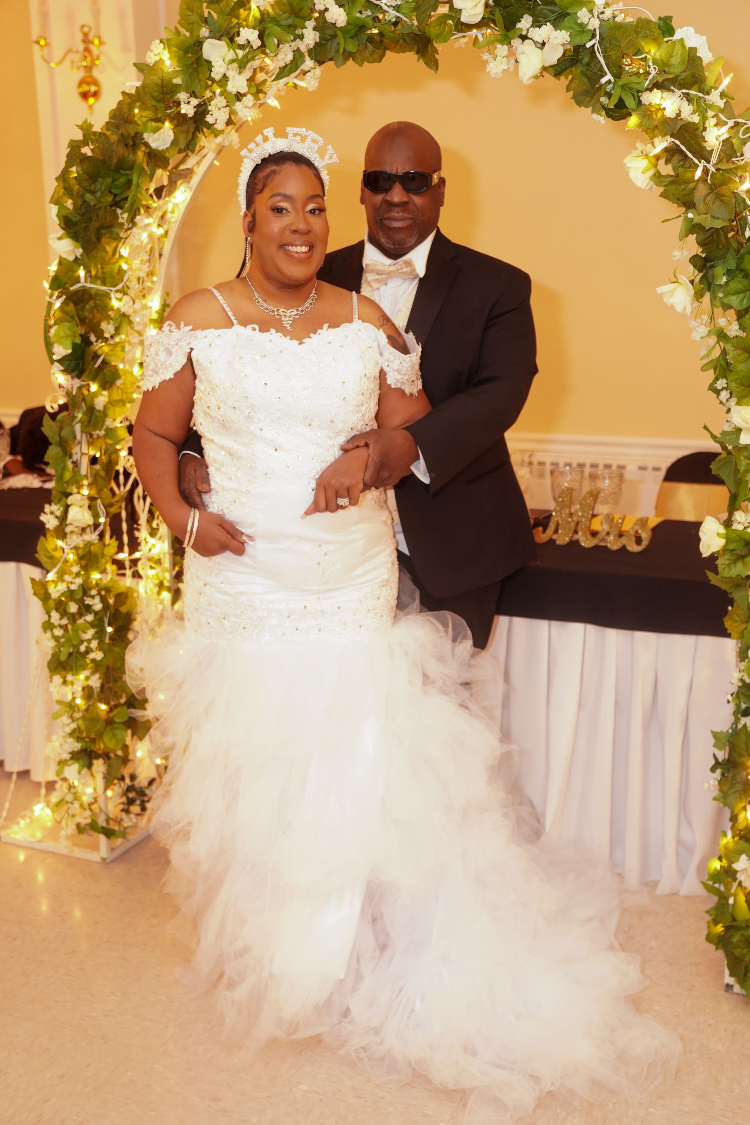 A couple poses under an elegant floral arch at their wedding, with the bride wearing a stunning white ruffled gown.