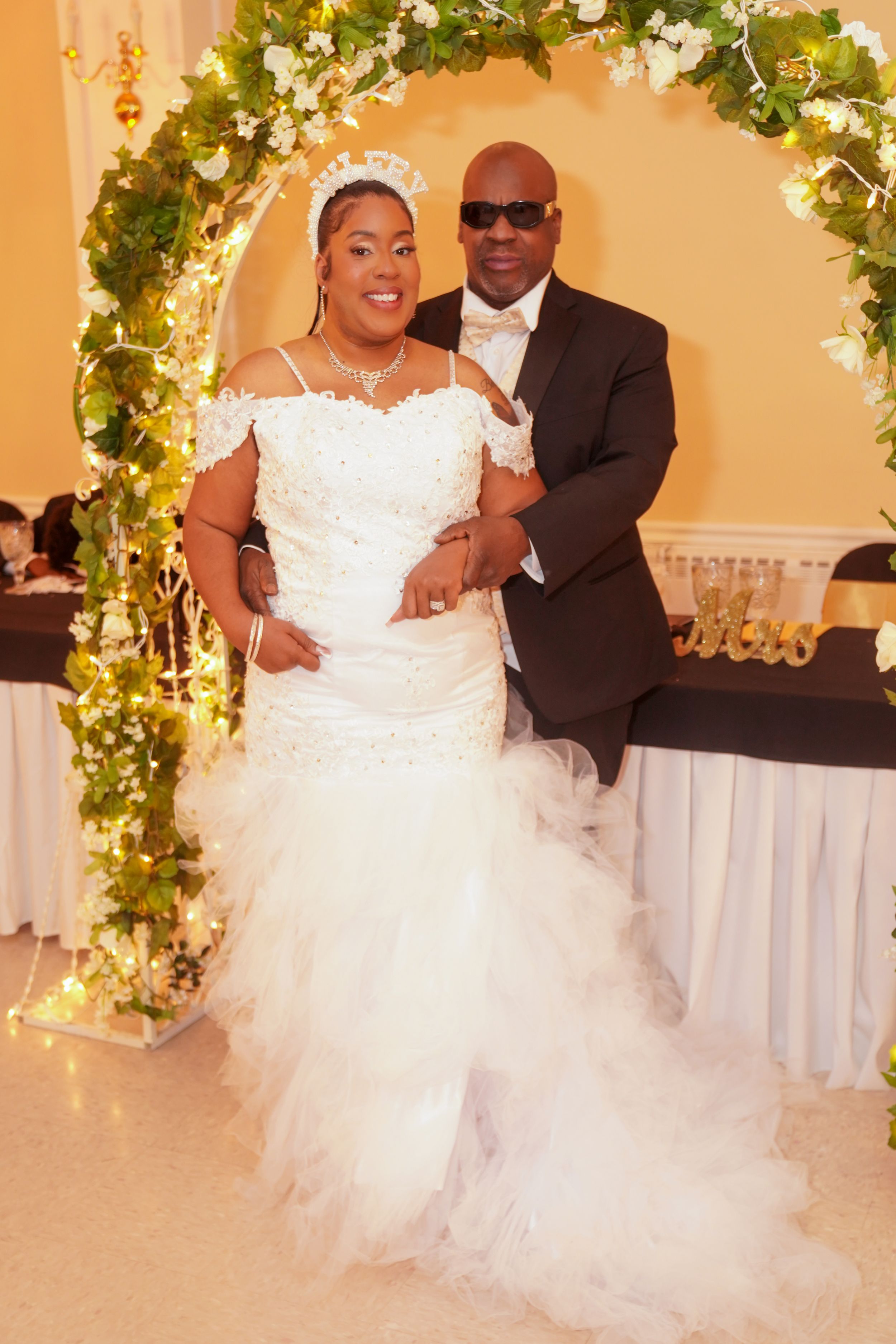 A couple in formal wedding attire poses under a floral arch during their reception.