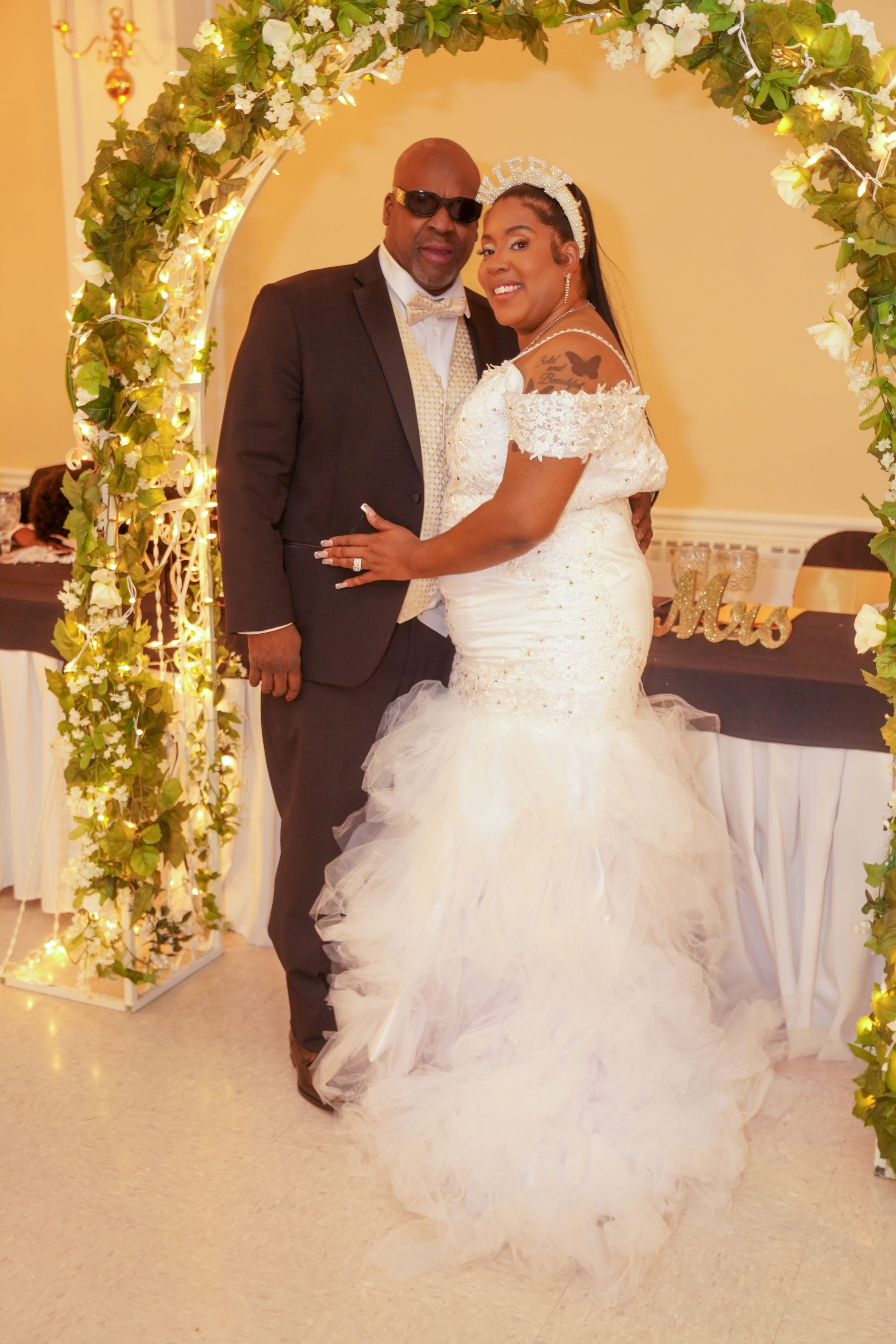 A couple poses under a beautiful floral wedding arch with twinkling lights at their elegant celebration.