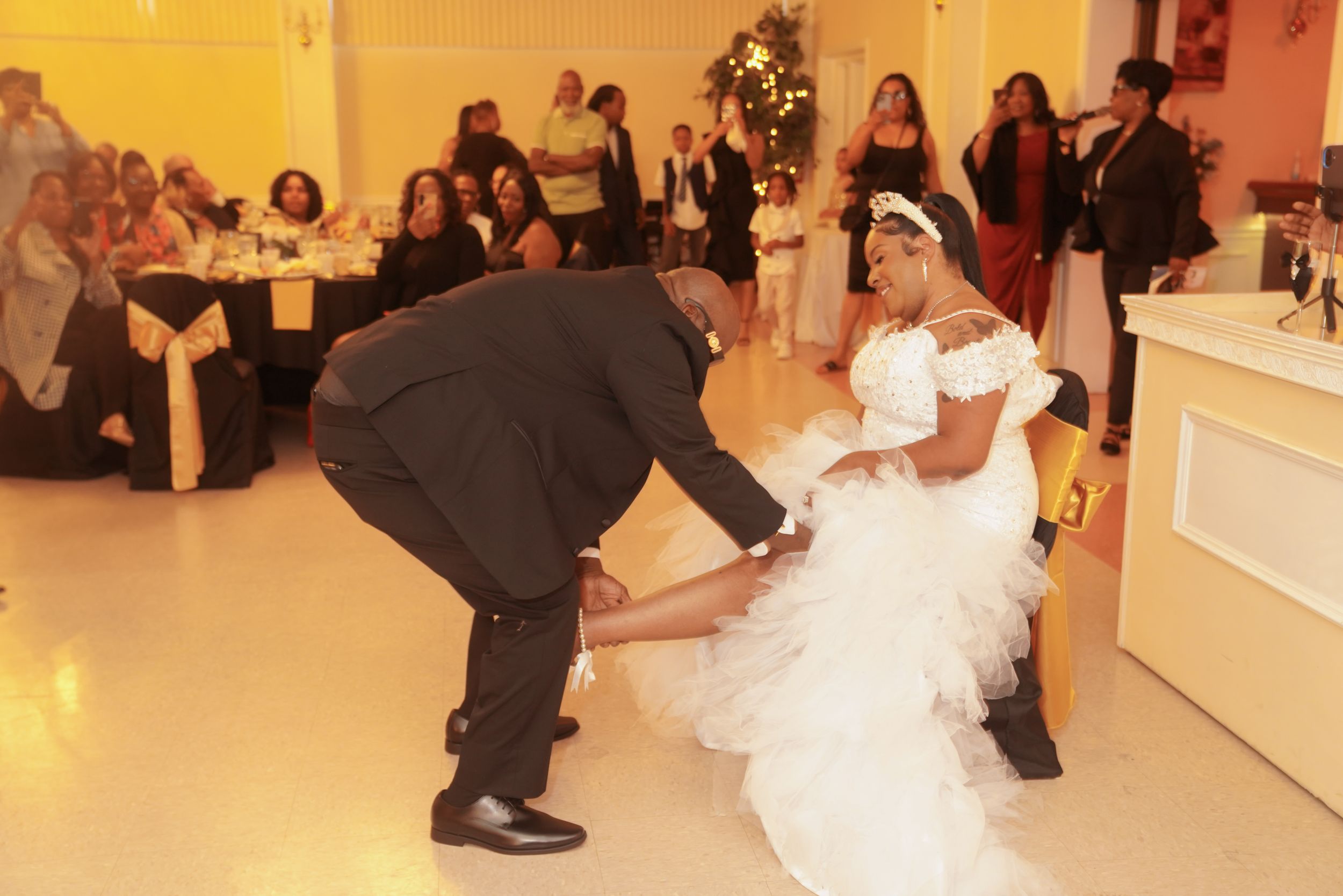Wedding guests watch as the groom helps adjust the bride's dress during a reception in an indoor venue.
