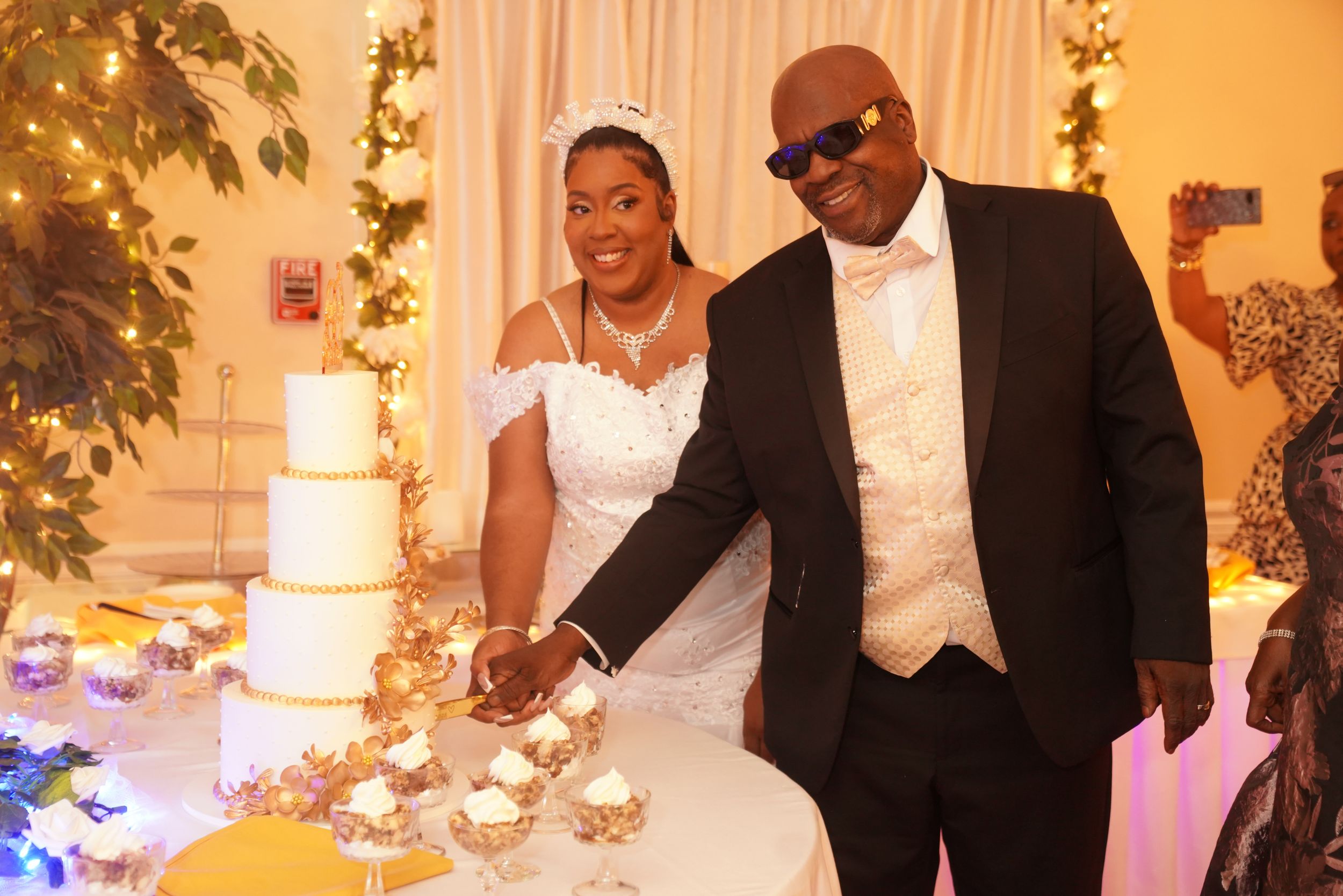 A happy couple cuts their elegant white wedding cake together at their reception with twinkling lights in the background.