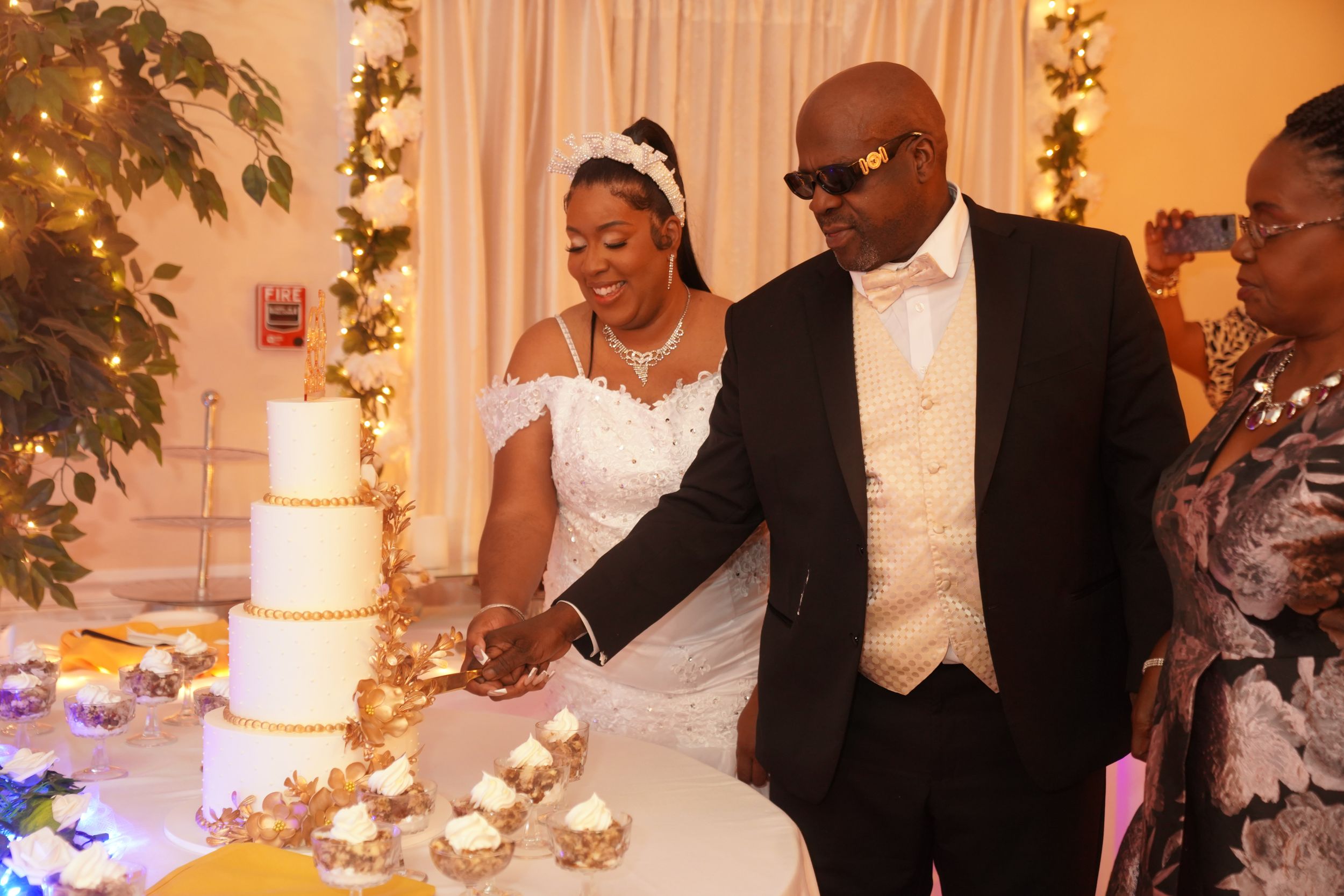 A couple cuts their elegant tiered wedding cake together at their reception while decorated with string lights and flowers.