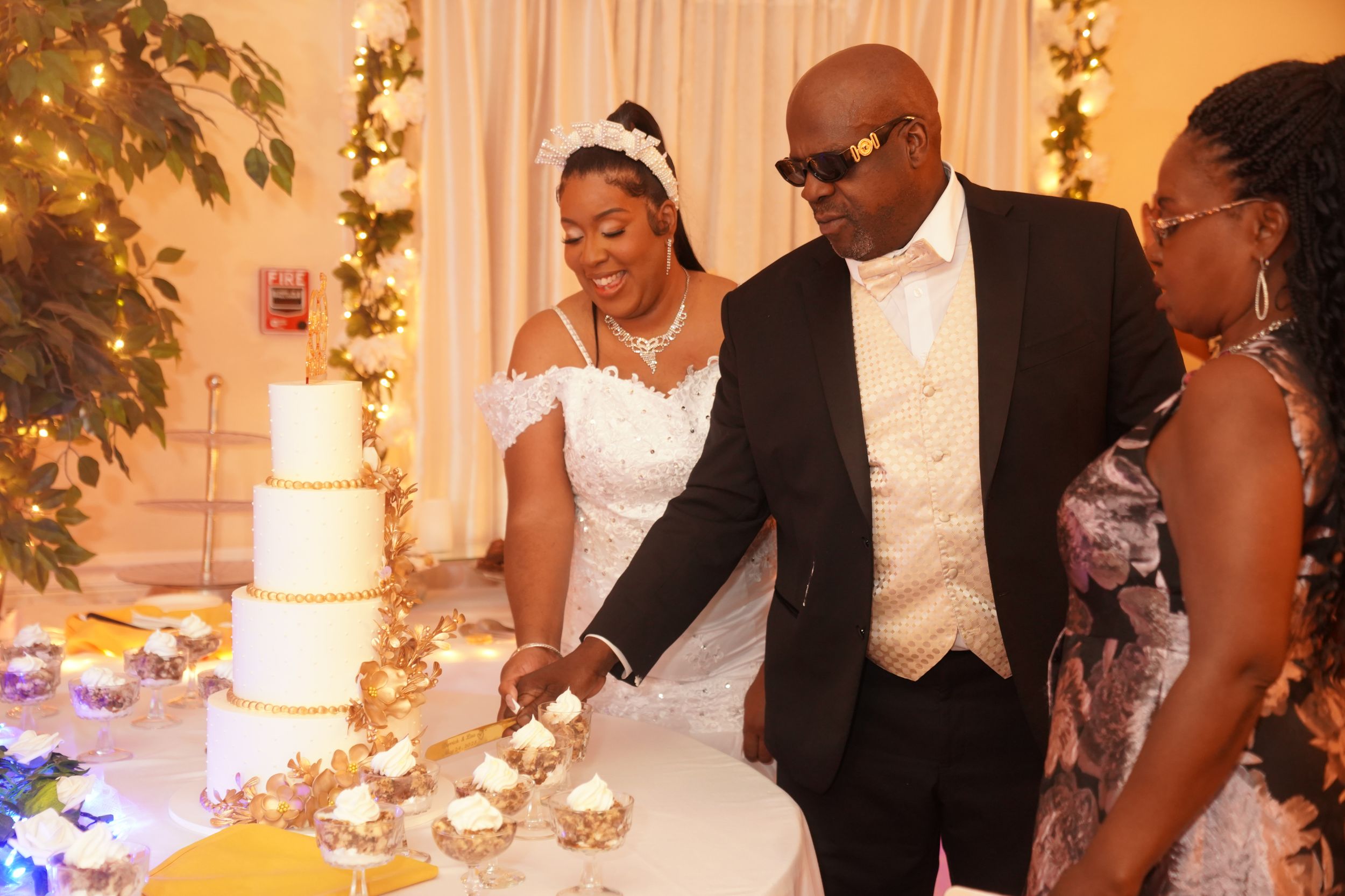 A newlywed couple and guest cutting their elegant tiered white wedding cake at the reception with decorative lights.