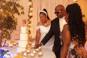 A wedding couple and guest slice into a multi-tiered white cake during an elegant evening reception ceremony.
