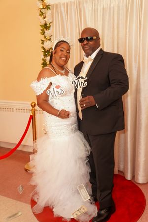 A formal wedding couple poses on a red carpet in front of a white backdrop with decorative vines.