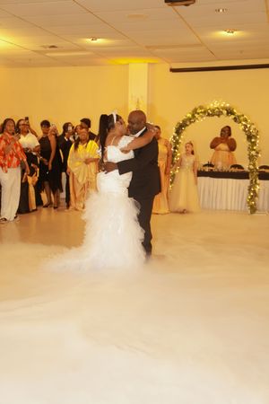 A wedding couple dances surrounded by guests near a floral arch in a mist-filled reception hall.