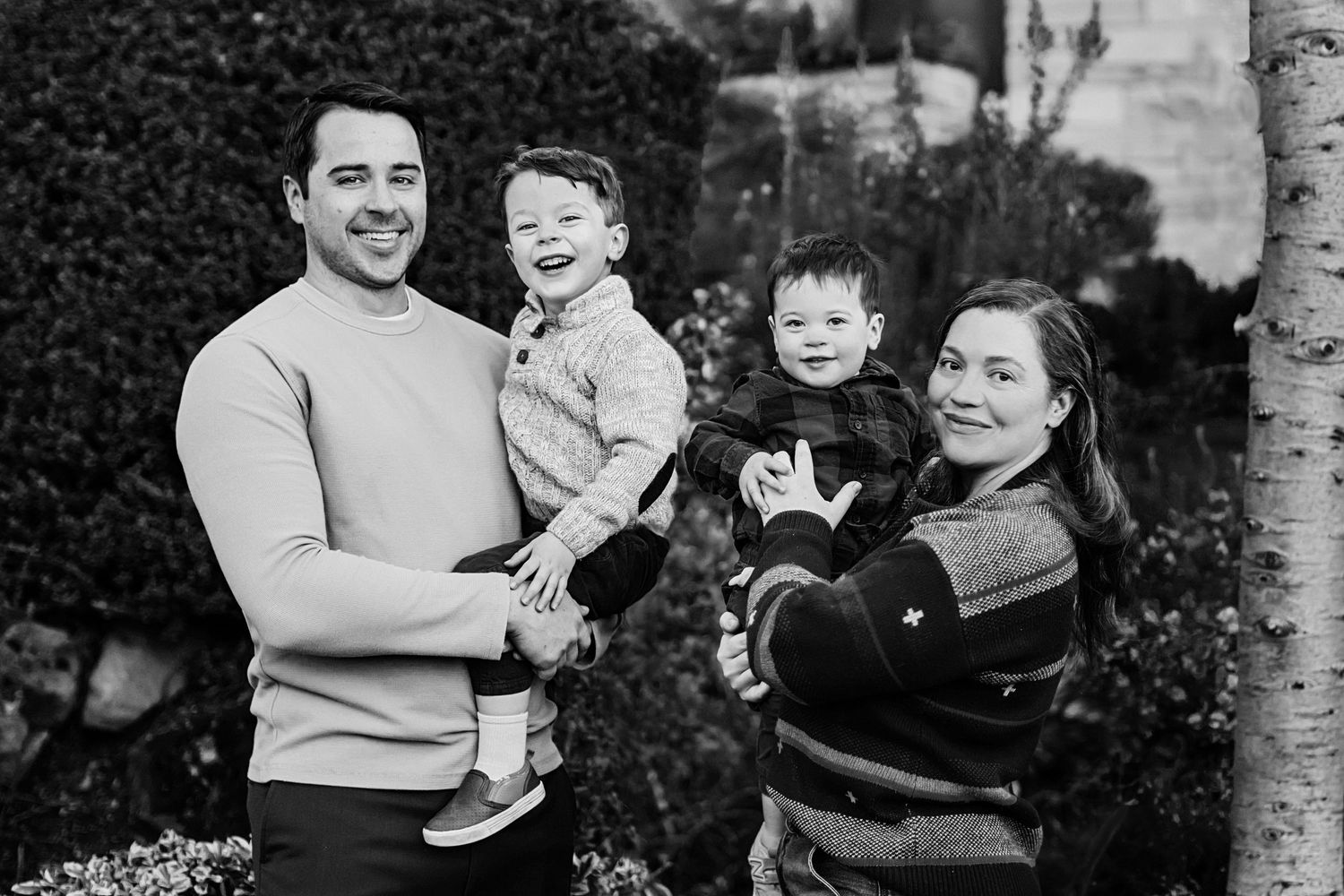 A four person family poses together outside in front of autumn foliage and green hedges.