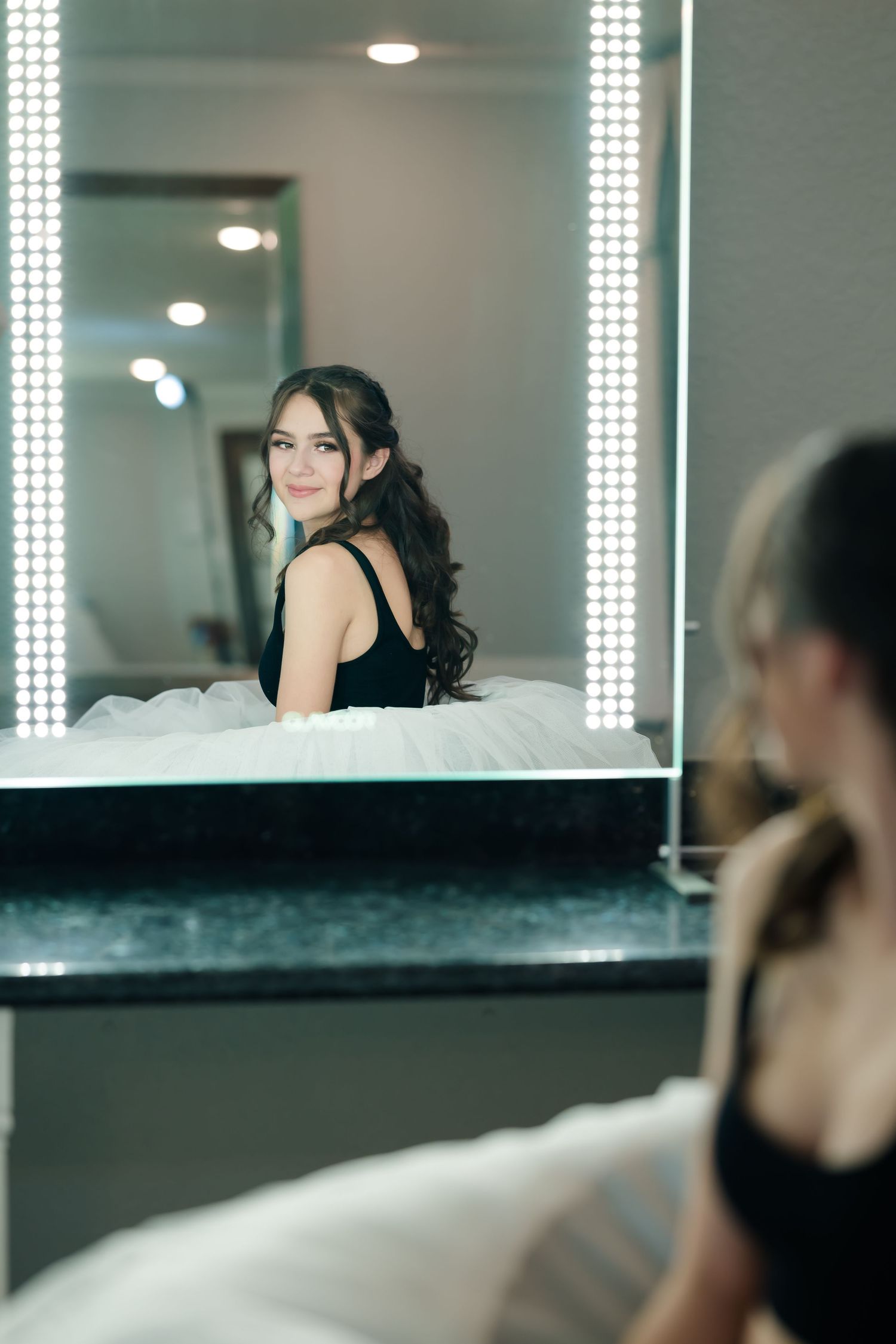 A reflection in a brightly lit vanity mirror shows an elegant figure in a black sleeveless dress.