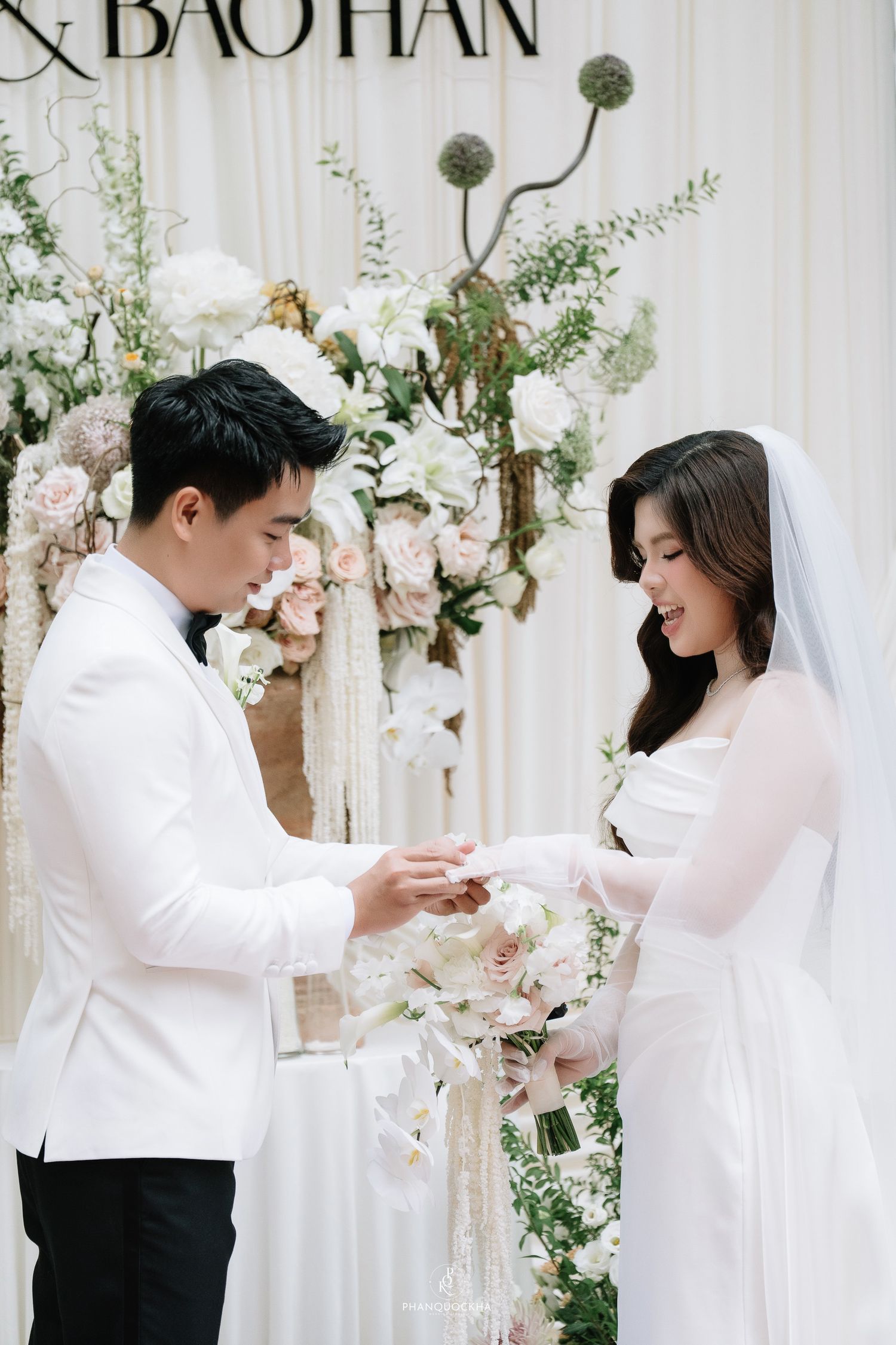 Groom places wedding ring on brides finger before lush floral arrangement at ceremony.