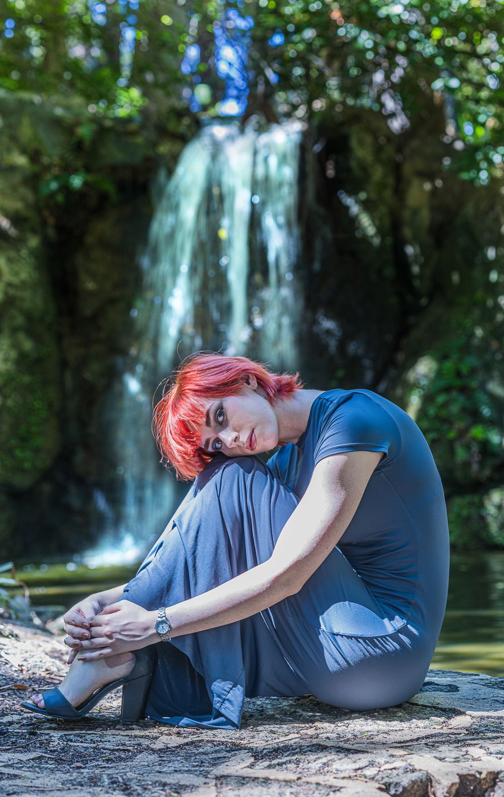 A woman relaxes on a rock by a waterfall, showcasing a serene moment during a model photoshoot in a scenic environment.